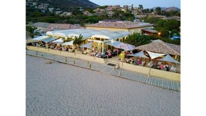A beachside restaurant with outdoor seating and umbrellas, overlooking a sandy beach and a hillside town. - Hotel Marina di Lava (Appietto)