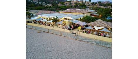 Un restaurante frente a la playa con terraza y sombrillas, con vista a una playa de arena y un pueblo en la ladera.