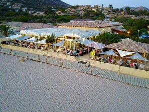 An outdoor seating area with umbrellas and tables, overlooking a beach and a hillside village. - Hotel Marina di Lava (Appietto)