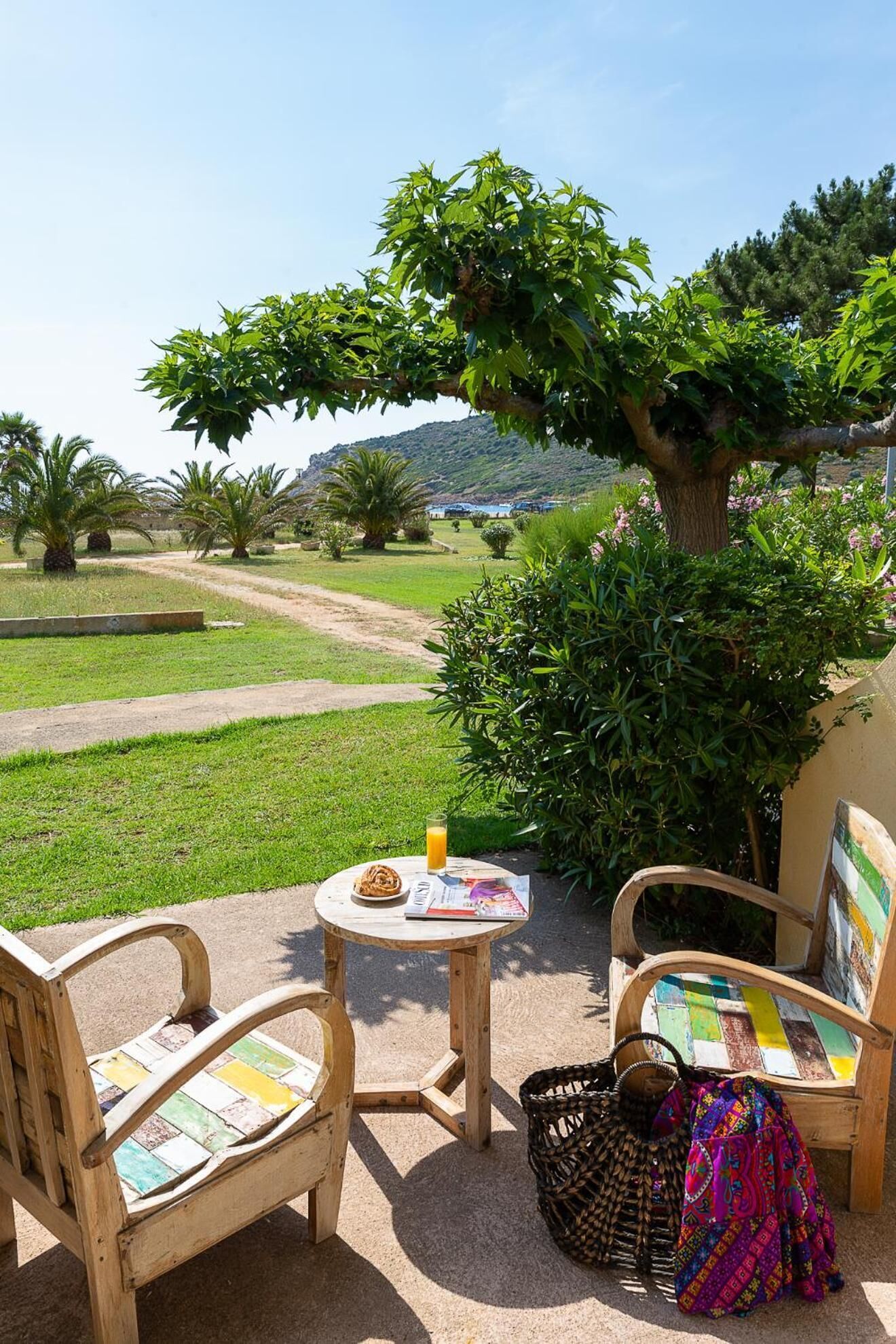 A patio with two wooden chairs, a small round table with a drink and a book, and a colorful woven bag.