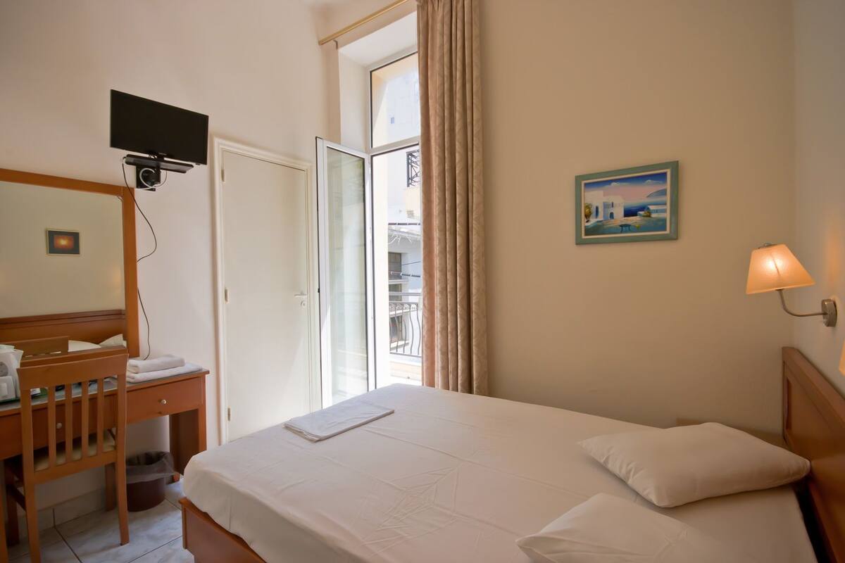 Habitación de hotel con cama, escritorio con silla, televisor montado en la pared, ventana con cortinas y un cuadro en la pared.