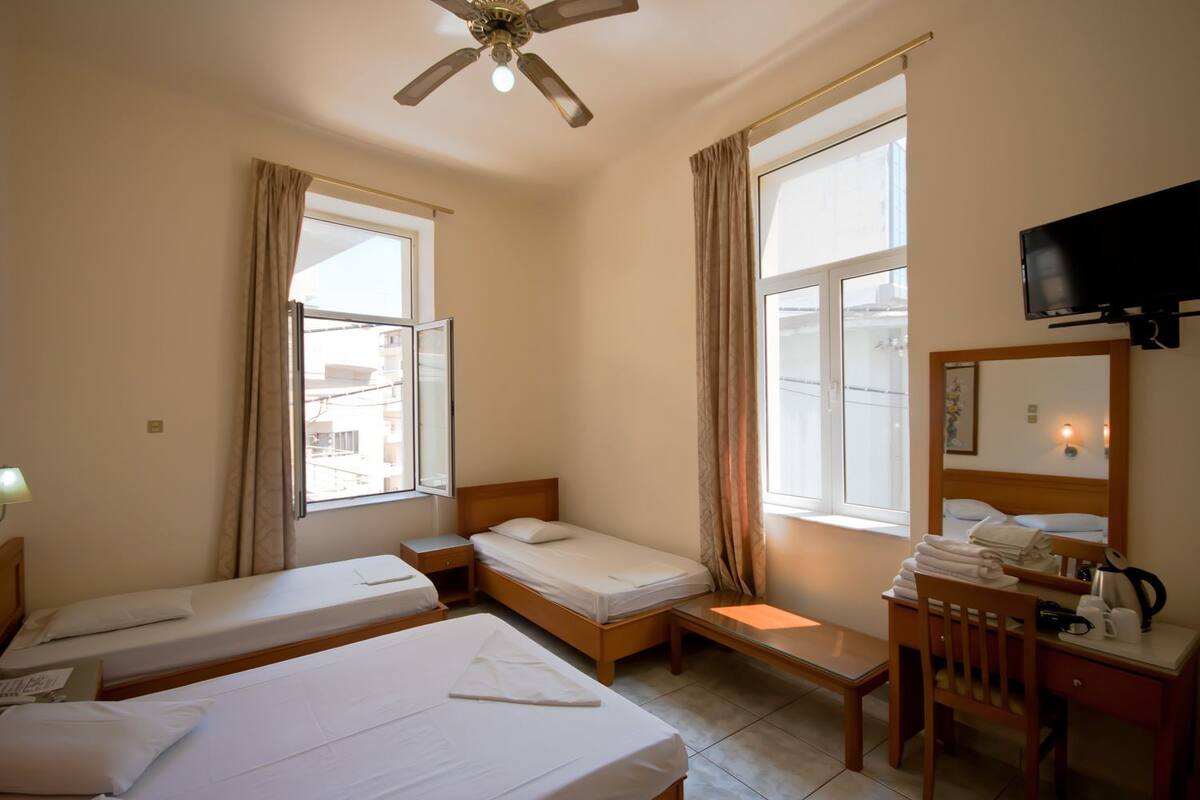 Habitación de hotel con dos camas individuales, un escritorio con silla, un televisor y un ventanal con cortinas.