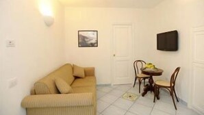 A small, well-lit living area with a beige sofa, a dining table with chairs, a framed picture on the wall, and a flat-screen TV mounted above the table. - Il Ducato di Ravello (Ravello)