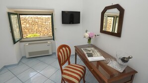 A room with a stone wall, a TV mounted on the wall, a wooden desk with a vase of flowers, a chair, and a window with an open door. - Il Ducato di Ravello (Ravello)