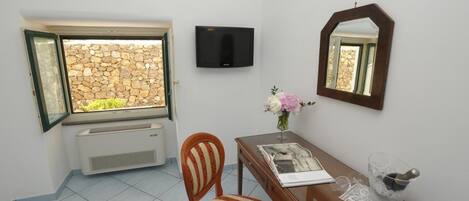 A room with a stone wall, a TV mounted on the wall, a wooden desk with a vase of flowers, a chair, and a window with an open door.