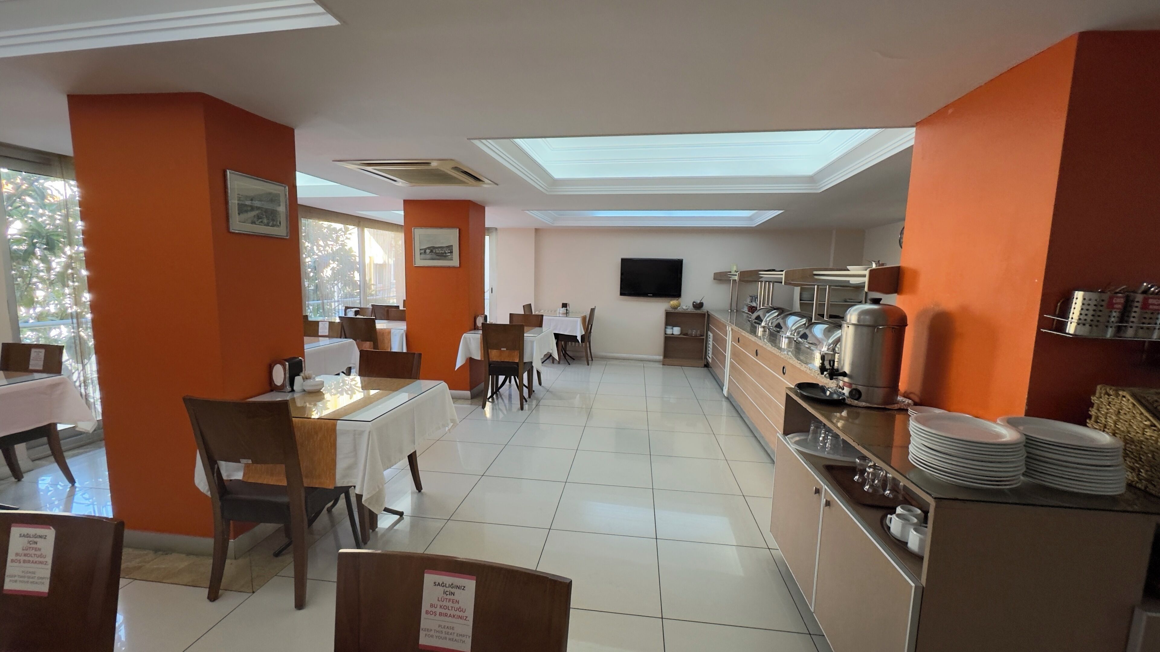 Un comedor con mesas y sillas, una barra de buffet con recipientes de comida y un televisor en la pared.