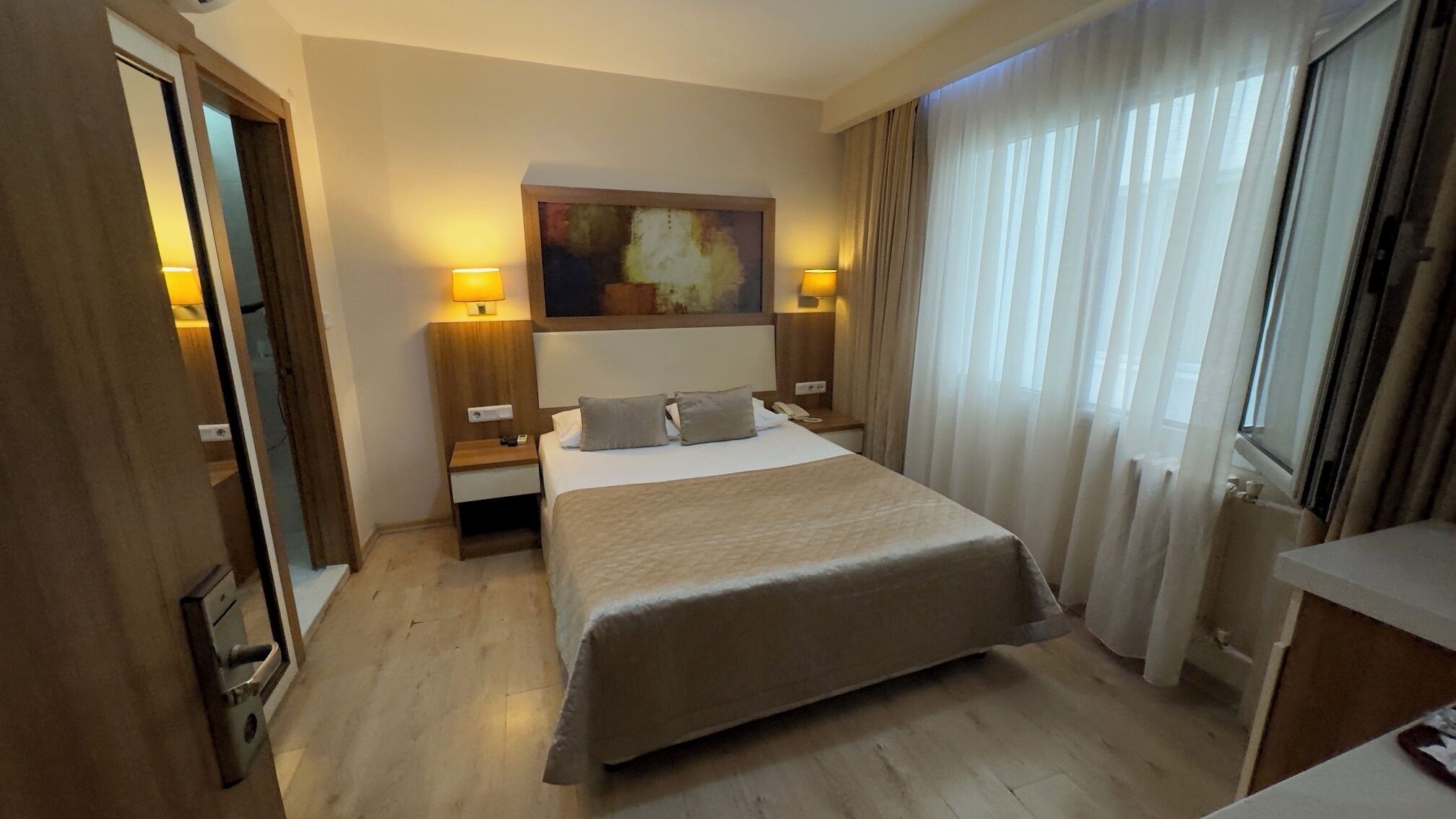 Una camera d'albergo con un letto, due comodini, un quadro appeso e una finestra con tende.