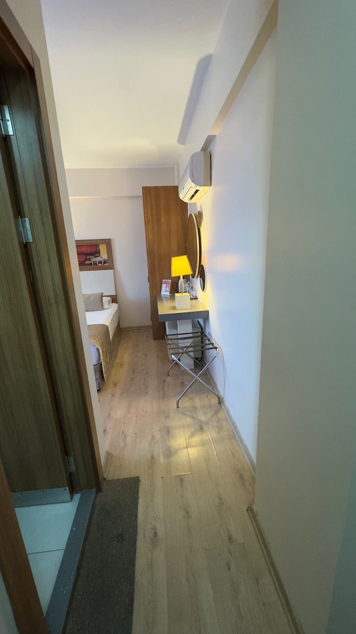 A narrow hallway with a wooden door on the left, a small desk with a lamp and a chair, and a wall-mounted air conditioner.