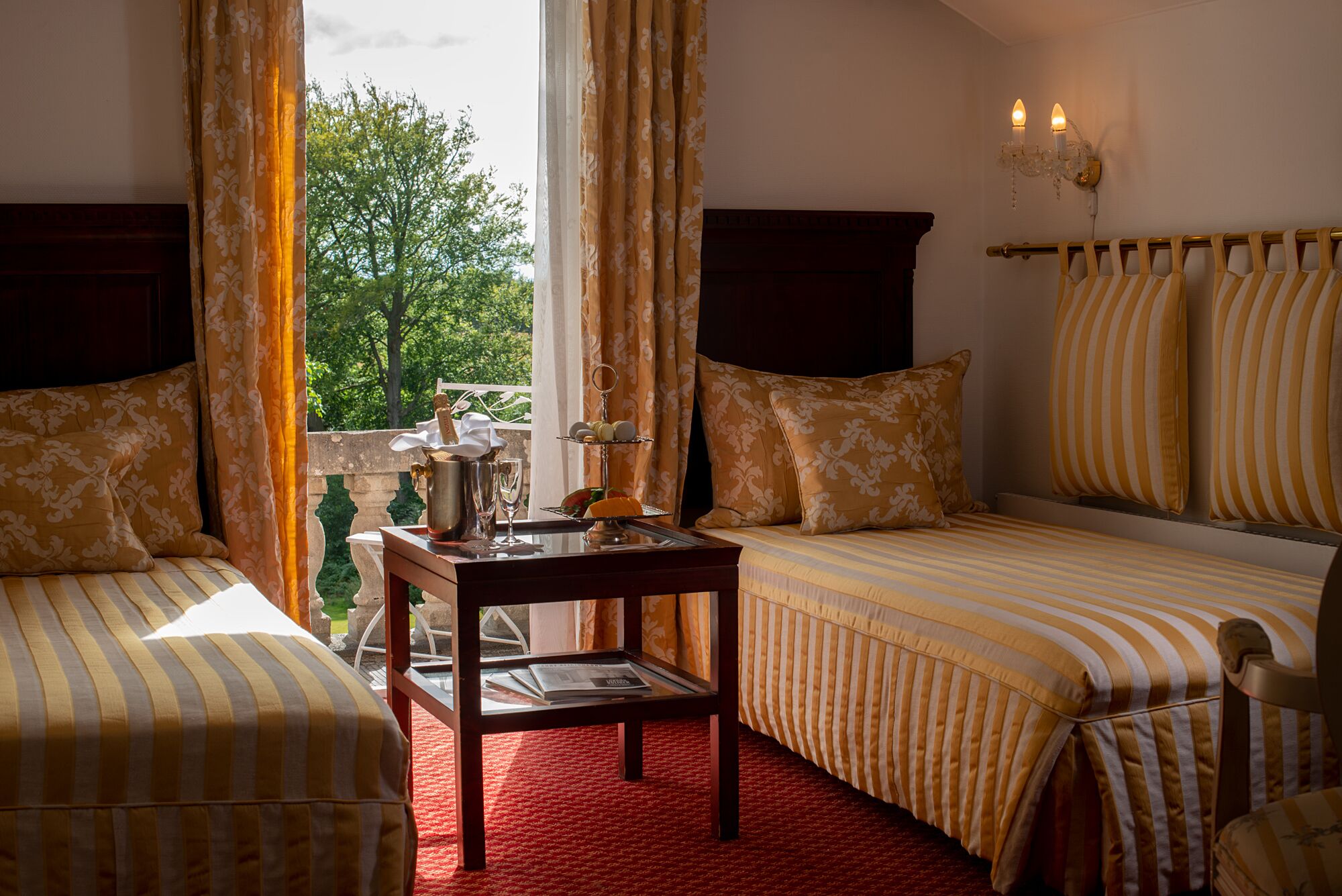 A room with two beds, a small table with a bottle and glass, and a view of trees through the window.