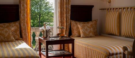 A room with two beds, a small table with a bottle and glass, and a view of trees through the window.