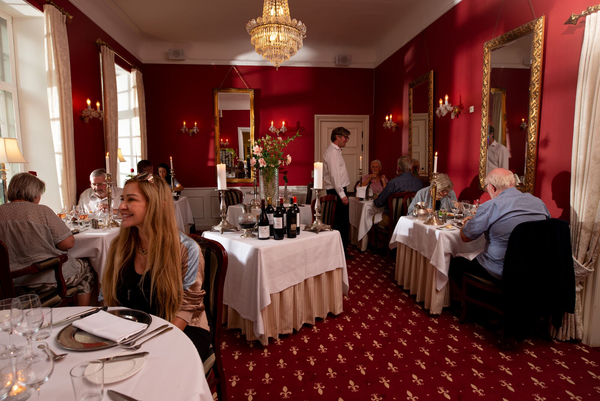 Comedor formal con paredes rojas, candelabros y espejos ornamentados. Las mesas están dispuestas con manteles blancos y velas.