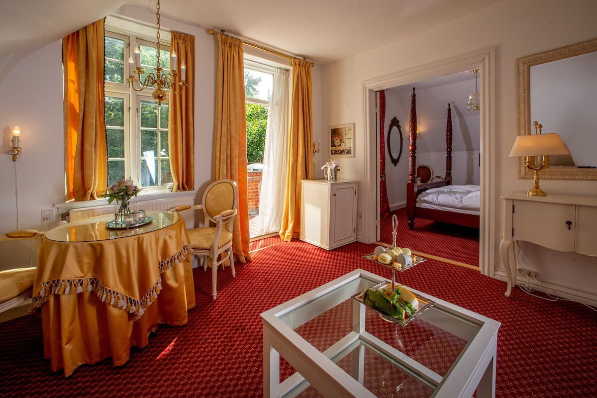 A hotel room with a red carpet, a dining area with a table and chairs, a bed with a canopy, and a mirror on the wall.