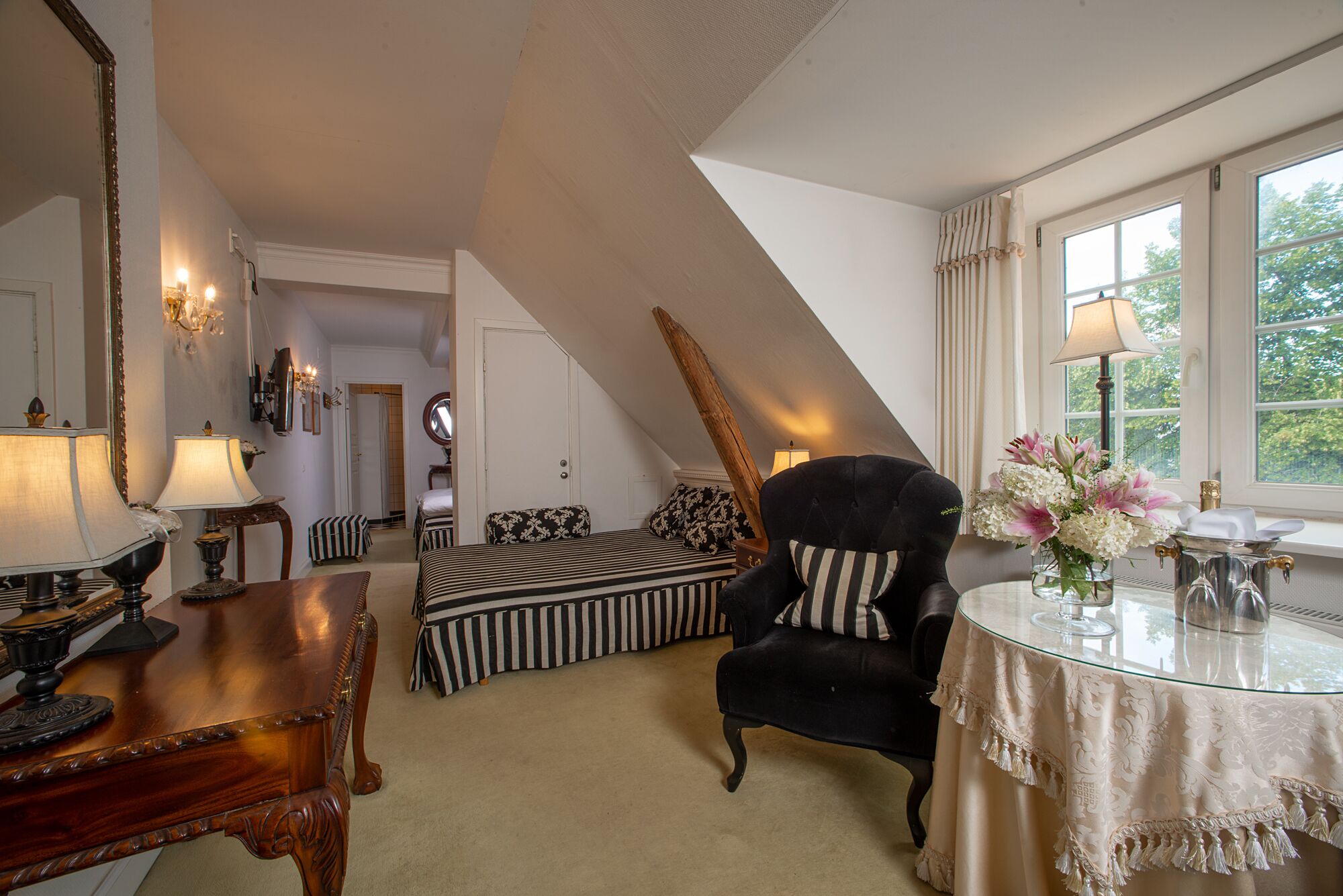 Een kamer met een hoge plafond, een zwarte fauteuil, een gestreepte bank, een houten tafel met glazen blad, en een raam met gordijnen.
