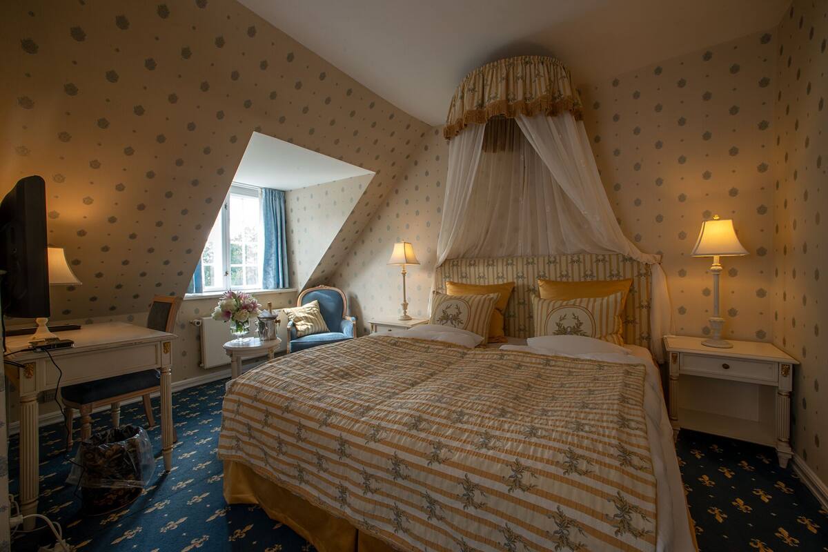 A bedroom with a large bed, two bedside tables with lamps, a chair, a desk with a computer, and a window with curtains.