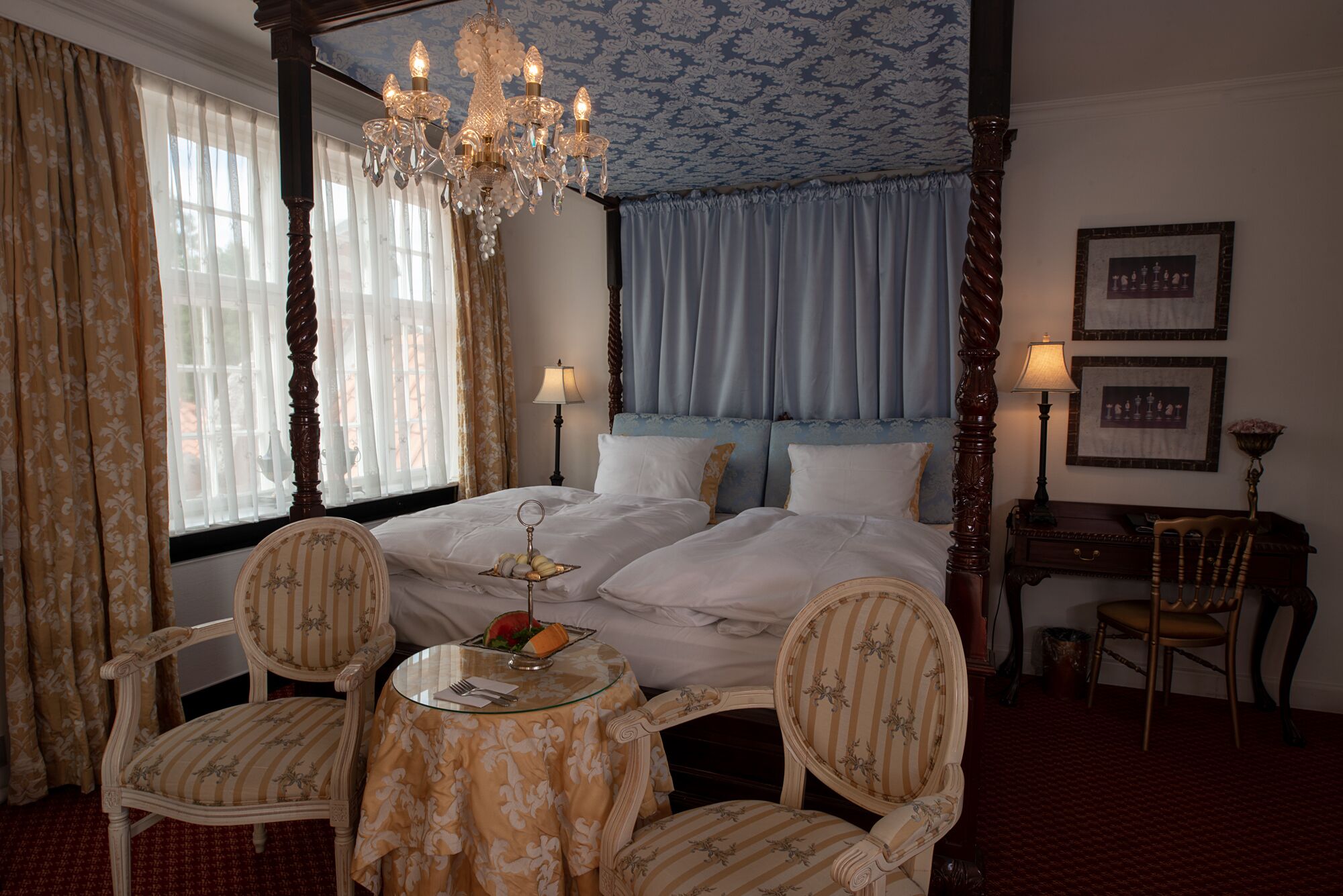 A four-poster bed with a chandelier, two upholstered chairs, a small round table with a glass top, and a desk with a lamp.