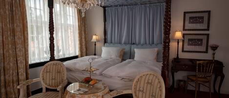 A four-poster bed with a chandelier, two upholstered chairs, a small round table with a glass top, and a desk with a lamp.