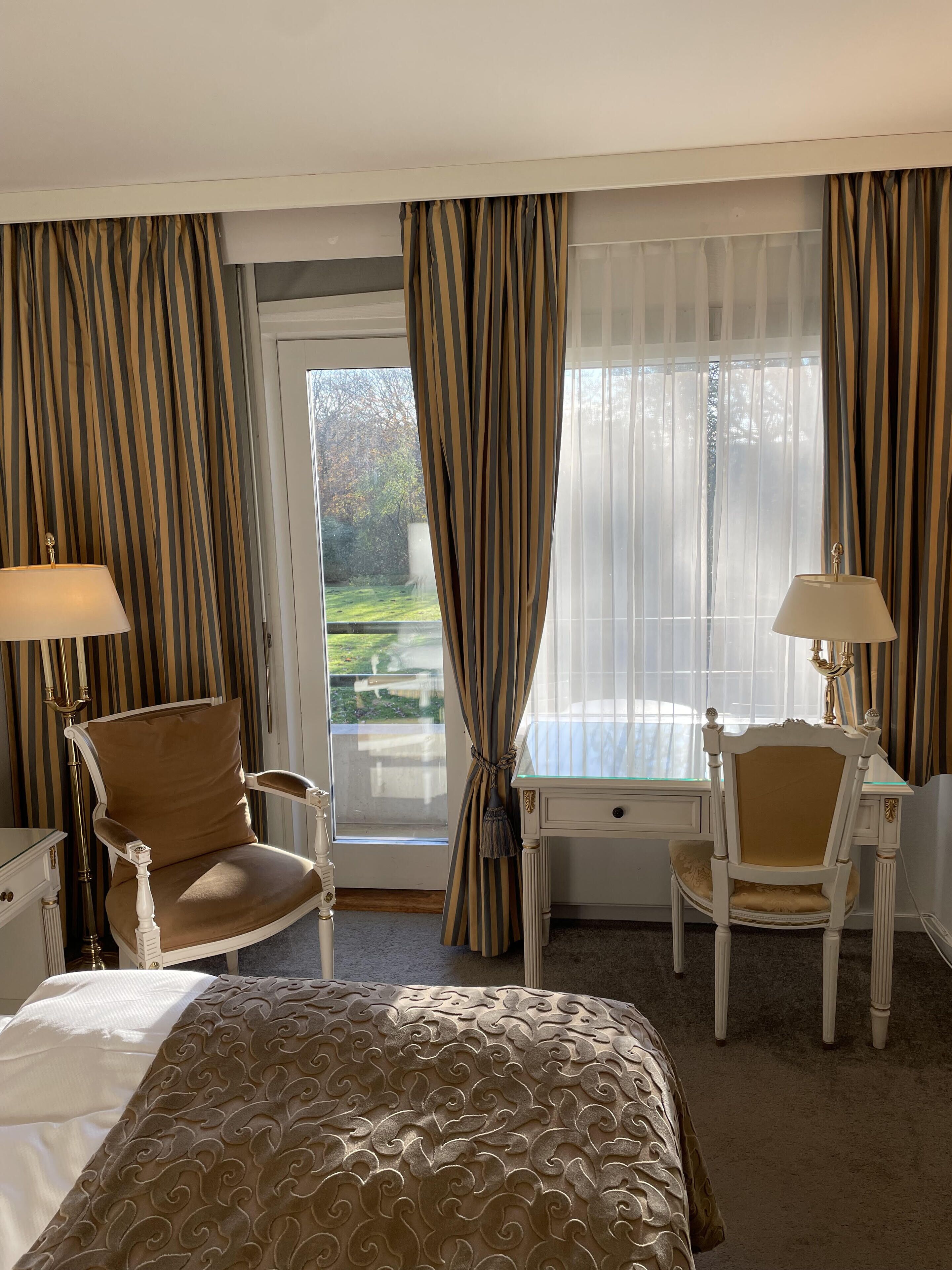 A hotel room with a bed, two chairs, a desk, and a view of a garden through the window.