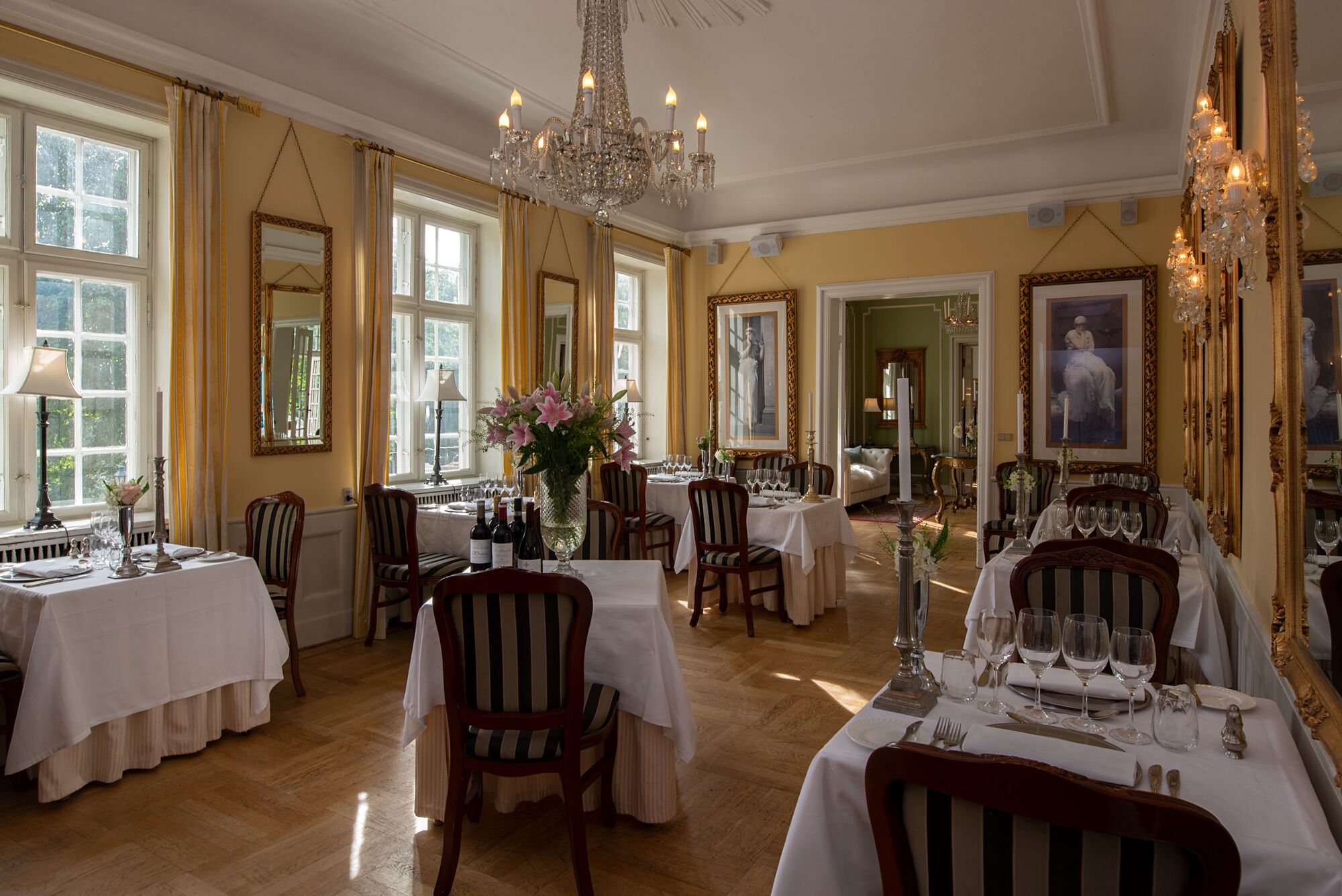 Un comedor elegante con varias mesas dispuestas para el servicio, ventanas amplias y candelabros clásicos.