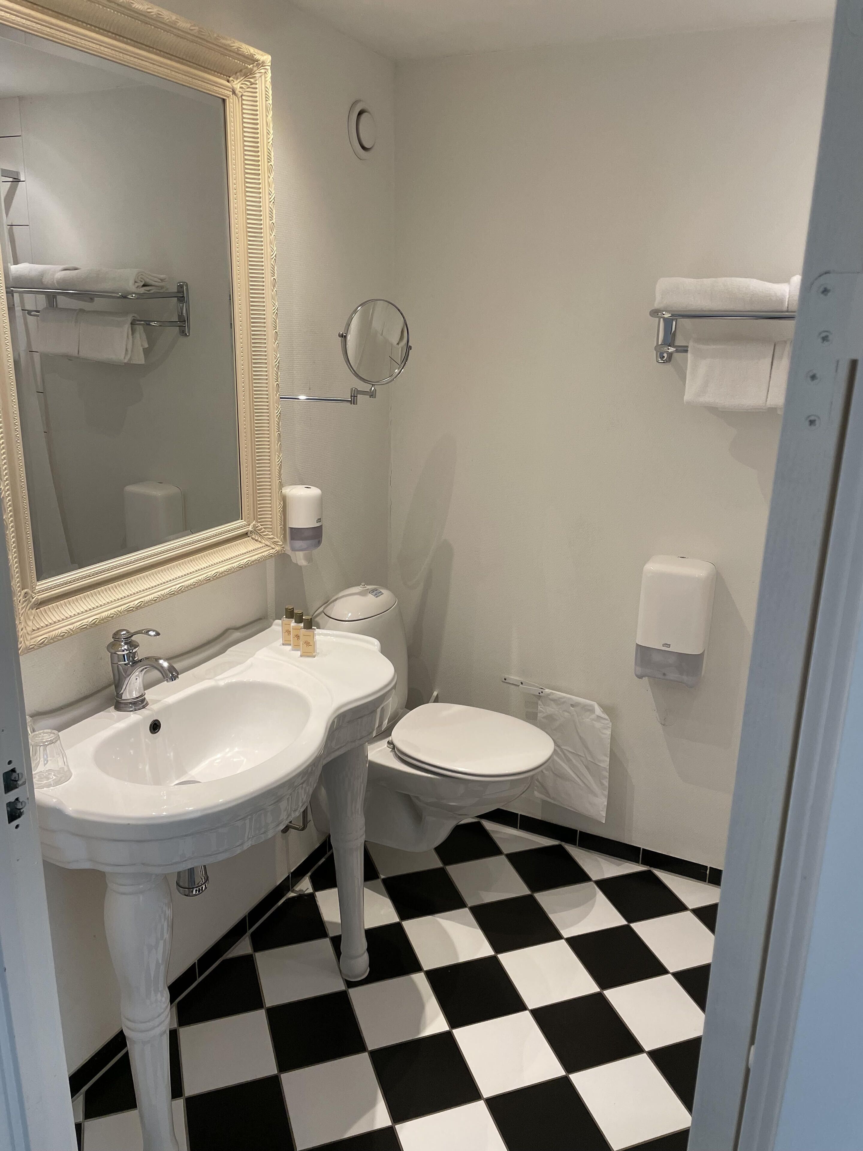 A bathroom with a white sink, a toilet, and a mirror with a decorative frame. There are two towel racks and a soap dispenser on the wall.