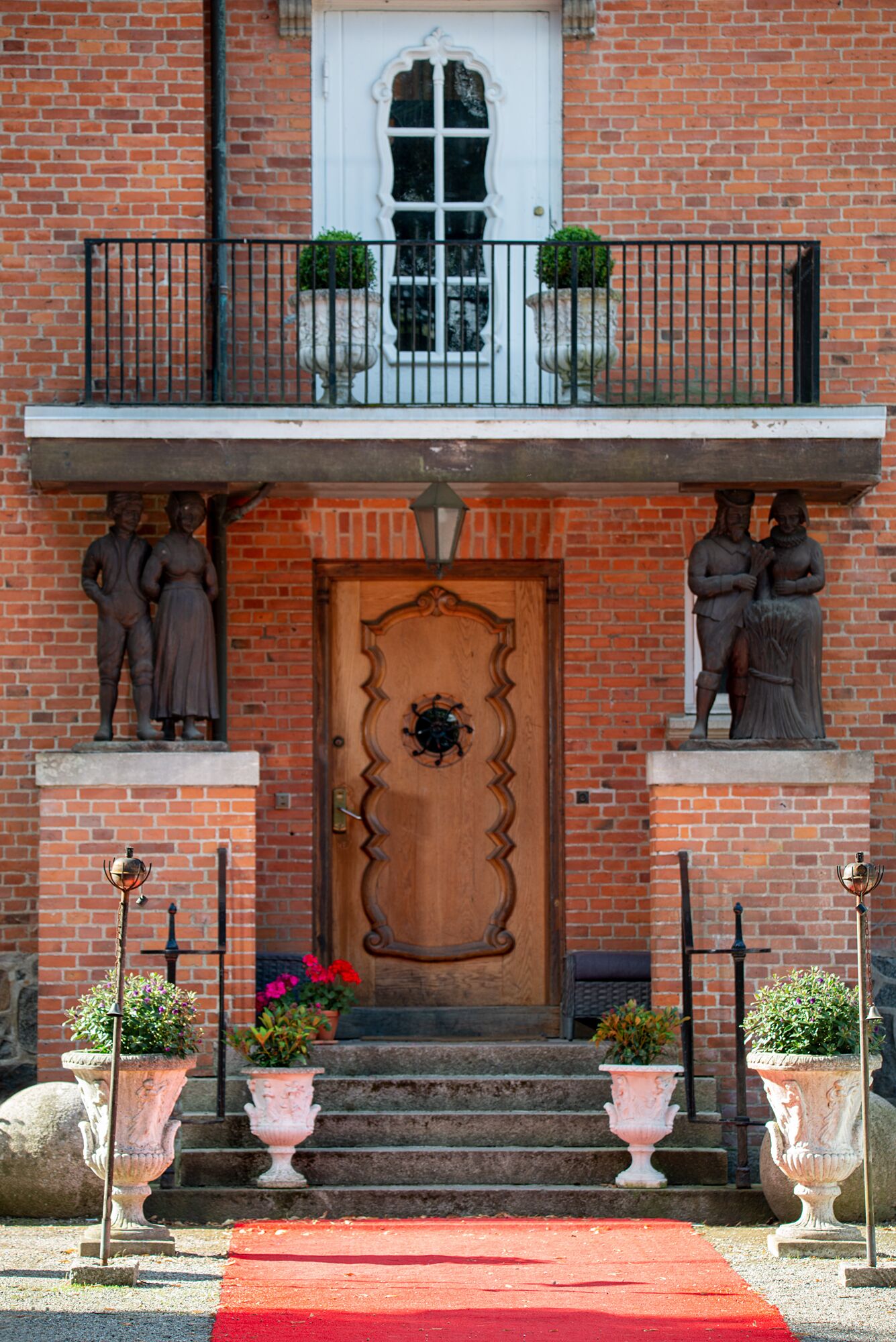 Un bâtiment en briques avec une porte en bois, un balcon doté de balustrades et deux statues de chaque côté de la porte.