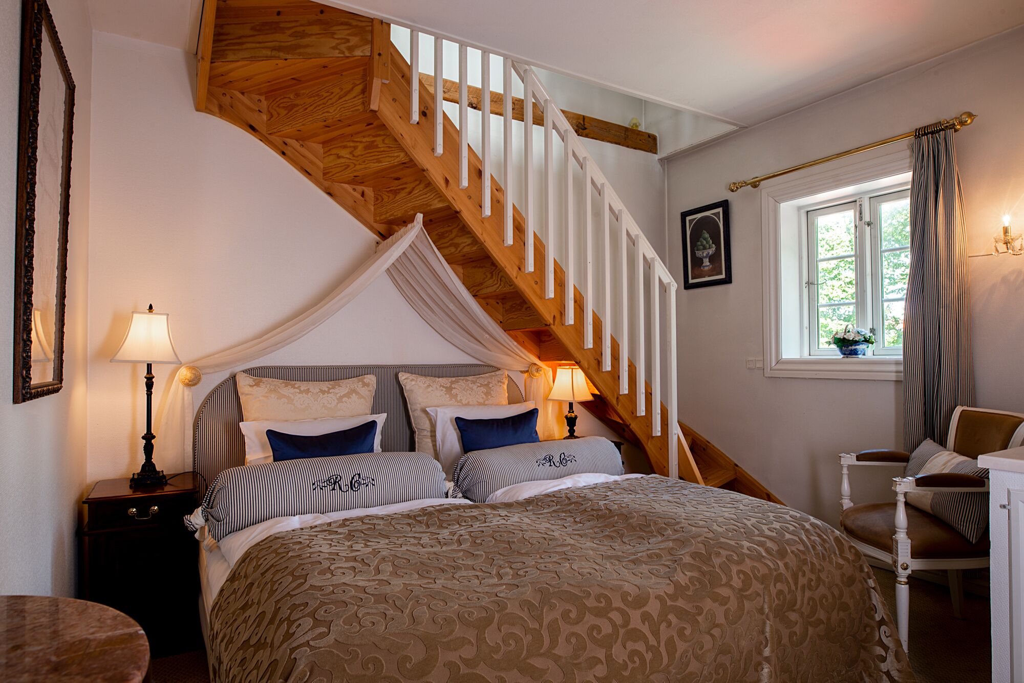 A bedroom with a wooden staircase, a canopy bed, bedside lamps, a window with curtains, and a chair.