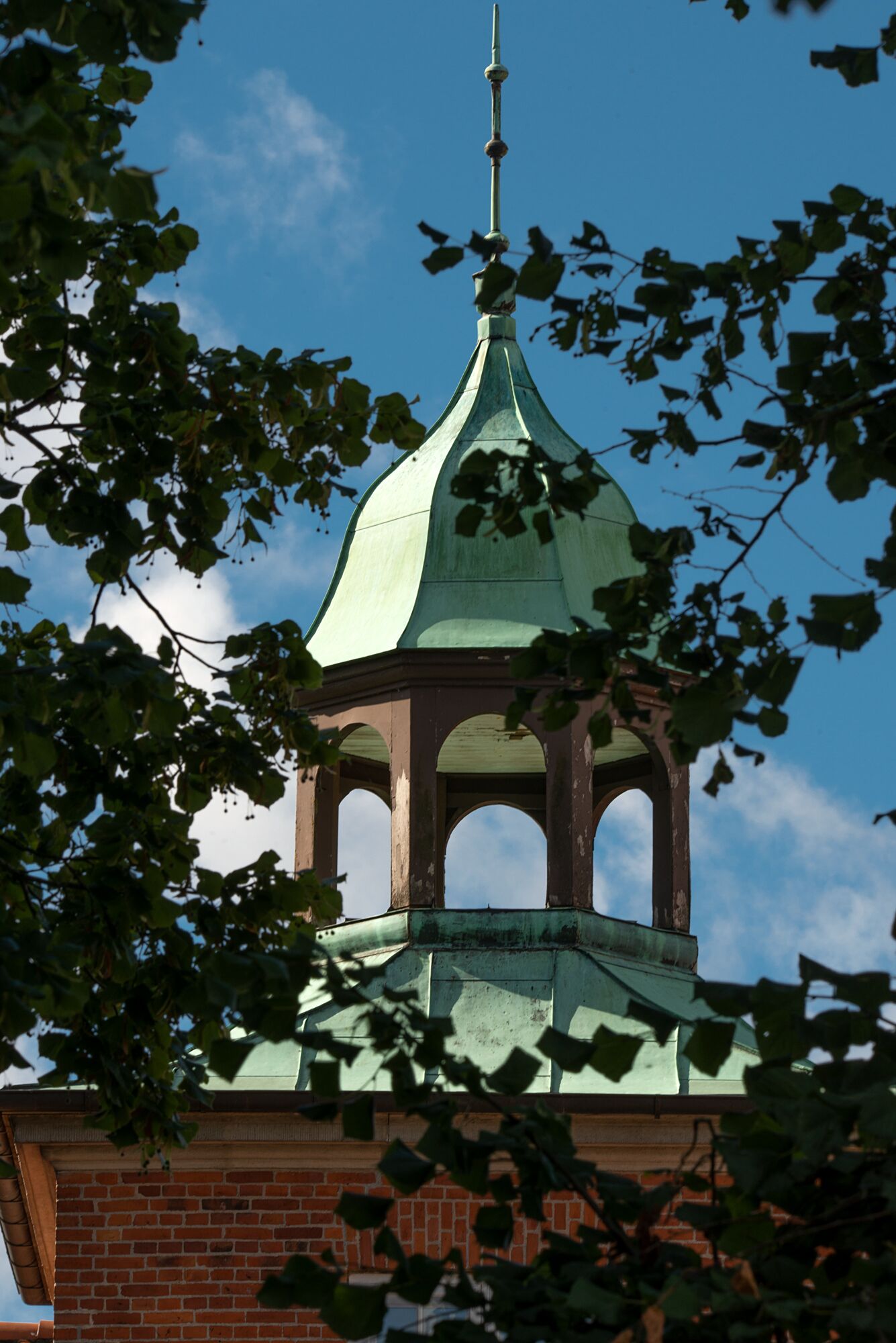 A brick building with a green-tiled bell tower and arched openings.