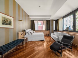 A modern hotel room with a wooden floor, a bed, a sofa, a bench, and a chair. There is a window with a view of buildings and a picture on the wall.