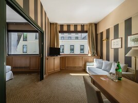 Una camera d'albergo con un'ampia finestra, una televisione su un mobile in legno, un divano, un tavolo con bicchieri e una bottiglia, e pareti a righe.