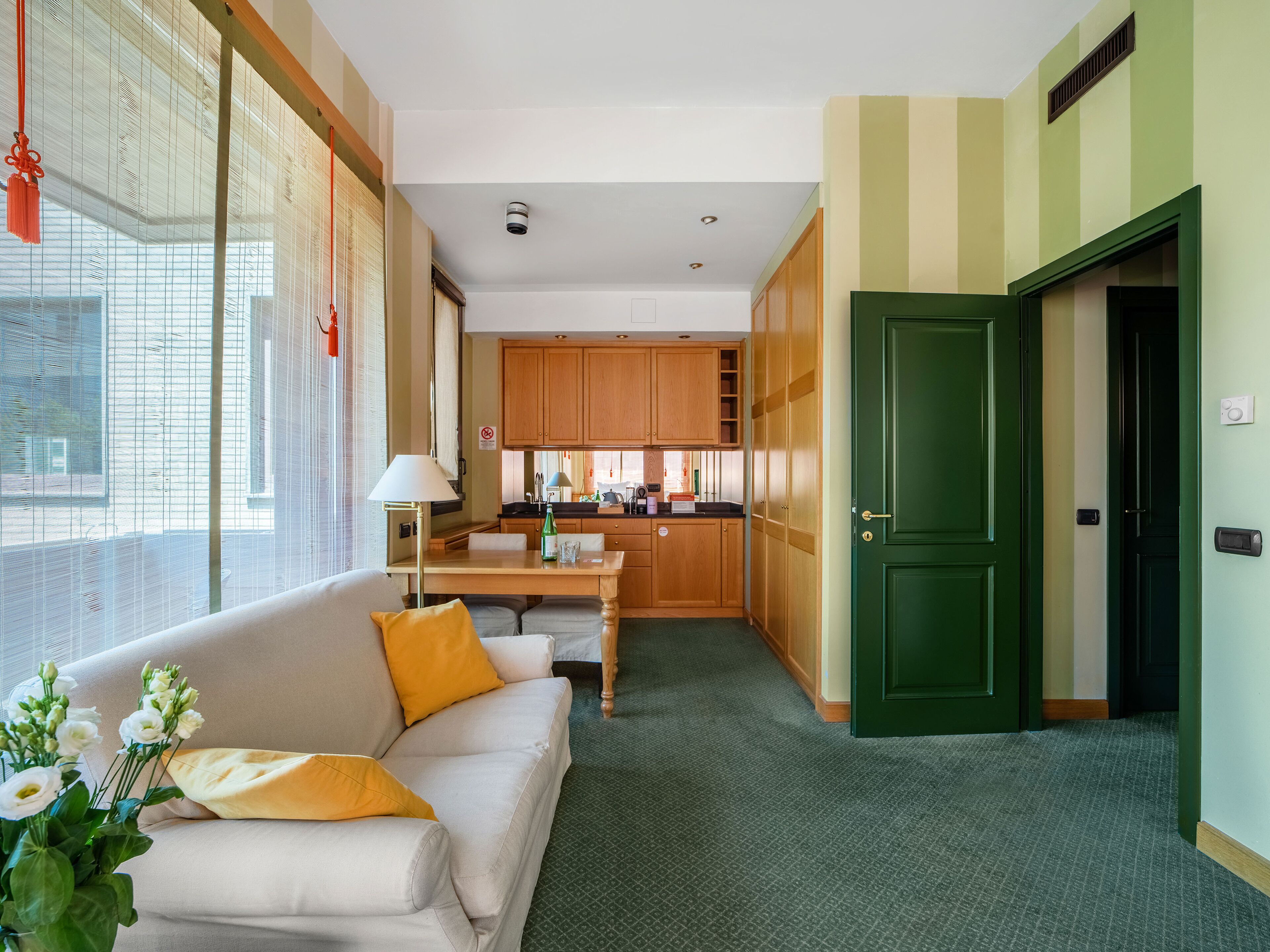 A hotel room with a sofa, a dining table, a kitchen area, and a green door.