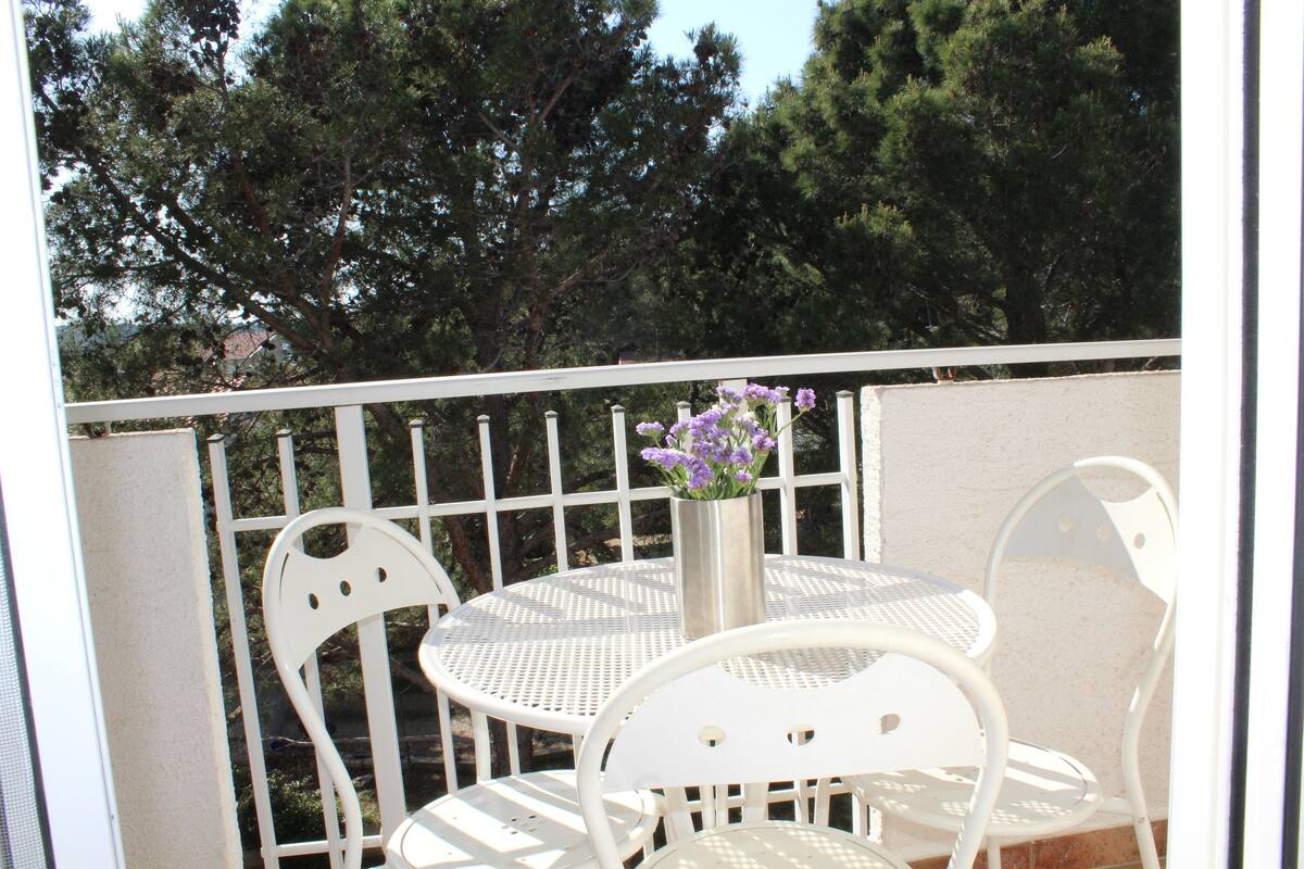 Un balcon avec une table ronde et deux chaises blanches, un vase rempli de fleurs violettes et une balustrade.