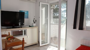 A room with a television on a cabinet, a dining table with chairs, a sliding glass door to the outside, and a bed with a red blanket.