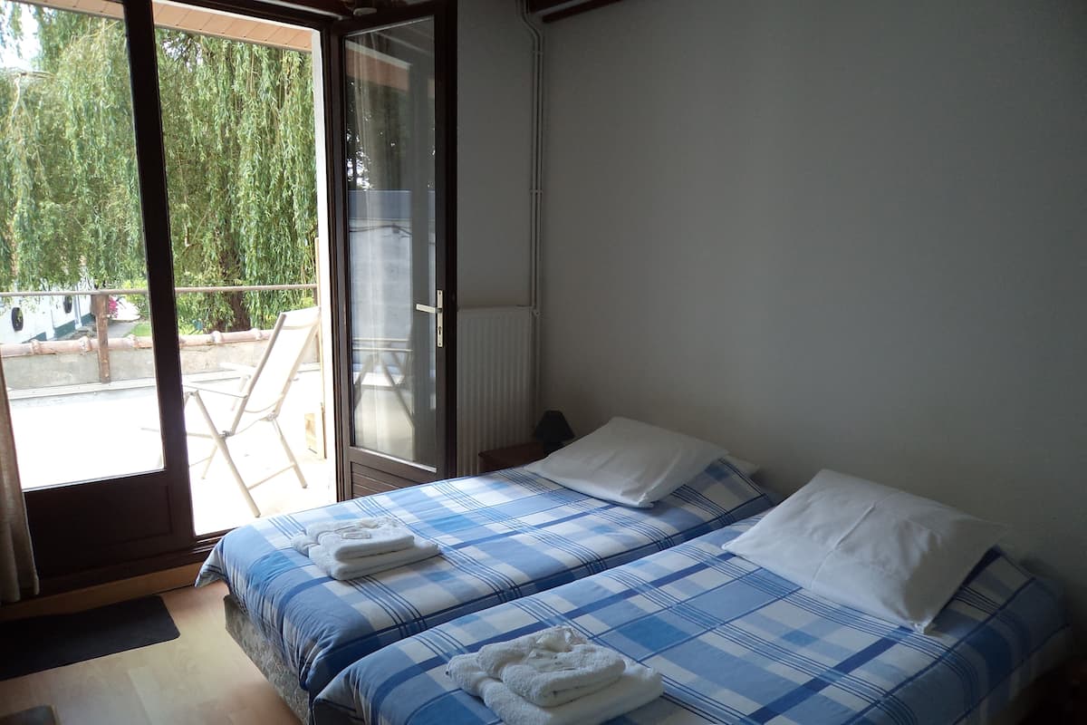 Un dormitorio con dos camas, cada una con una colcha a cuadros azules y blancos y almohadas blancas. Hay un ventanal corredera que da a un área exterior con una silla y una mesa.