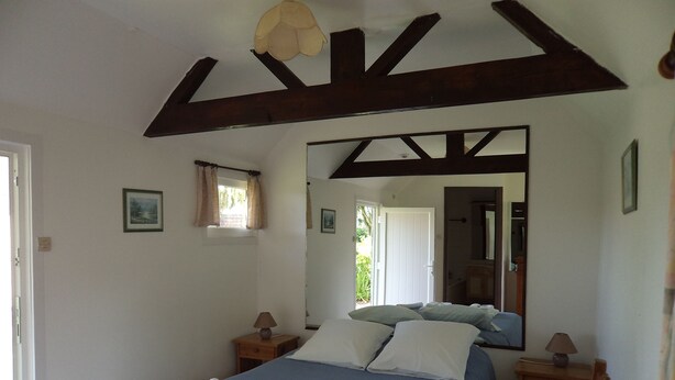 Een slaapkamer met een groot bed, houten balken, een raam met gordijnen, een nachtkastje, een spiegel en een radiator.