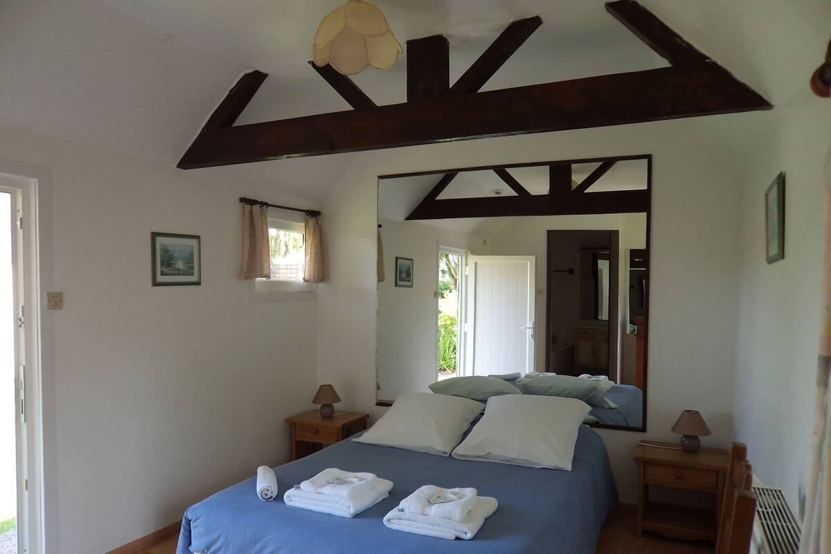 Un dormitorio con una cama grande, vigas de madera, una ventana con cortinas, una mesita de noche, un espejo y un radiador.