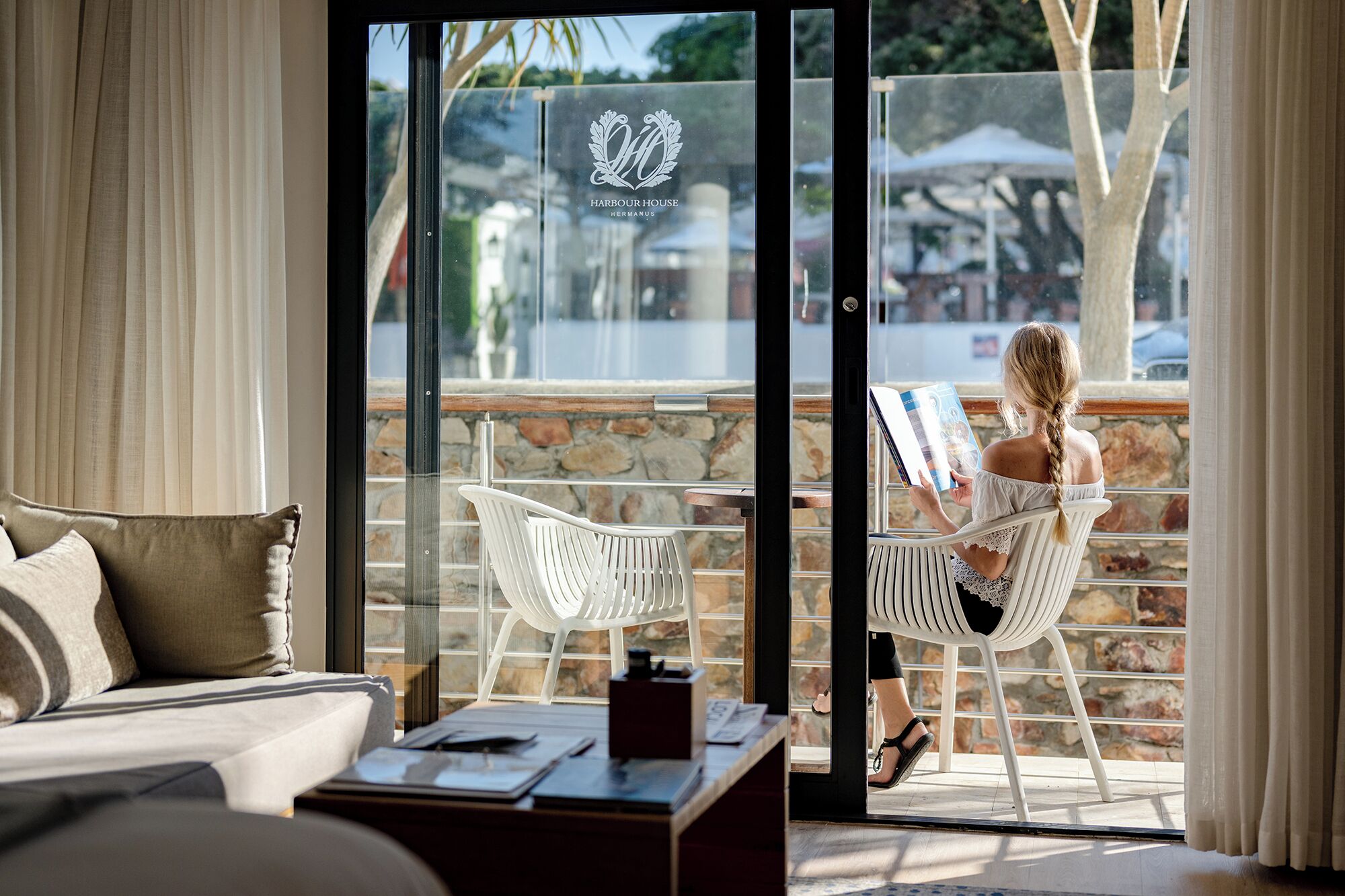 Ein Hotelzimmer mit einer Couch, einem Couchtisch und einer Glastür, die auf einen Balkon mit weißen Stühlen und einer Steinmauer führt.