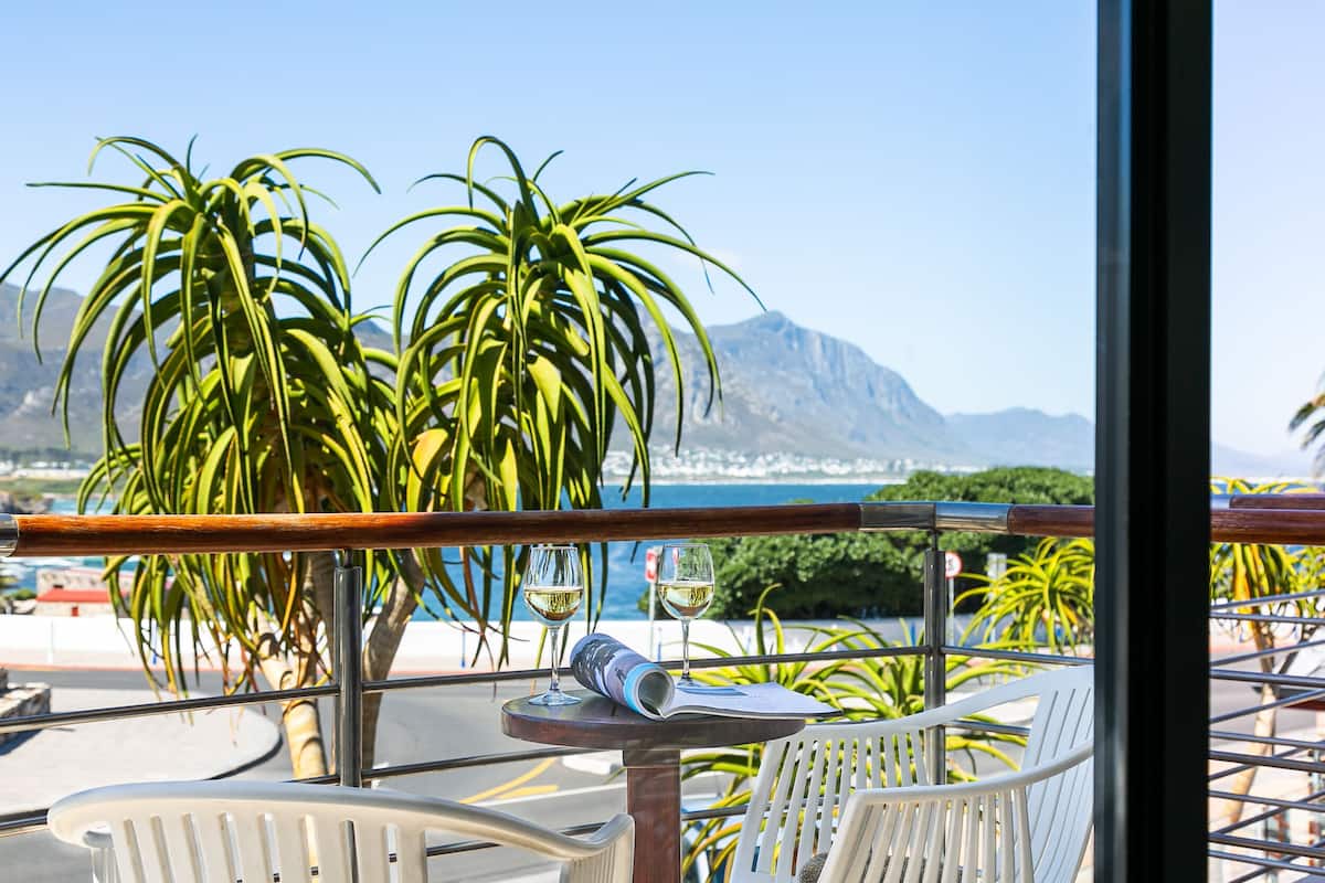 Una terraza con sillas blancas, una mesa con un menú enrollado y dos copas de vino blanco. Una baranda de madera ofrece vistas panorámicas del mar y las montañas.