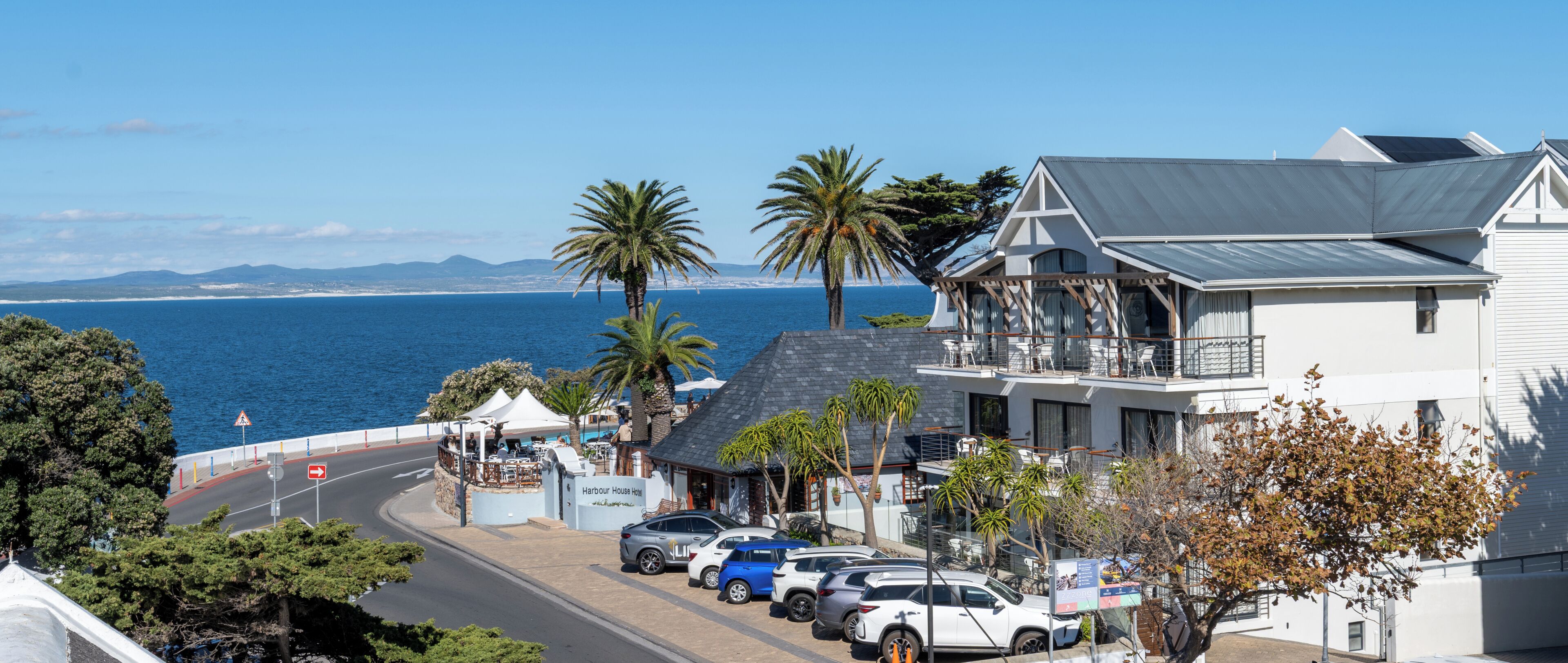 A coastal hotel with a balcony, parked cars, and a view of the sea.
