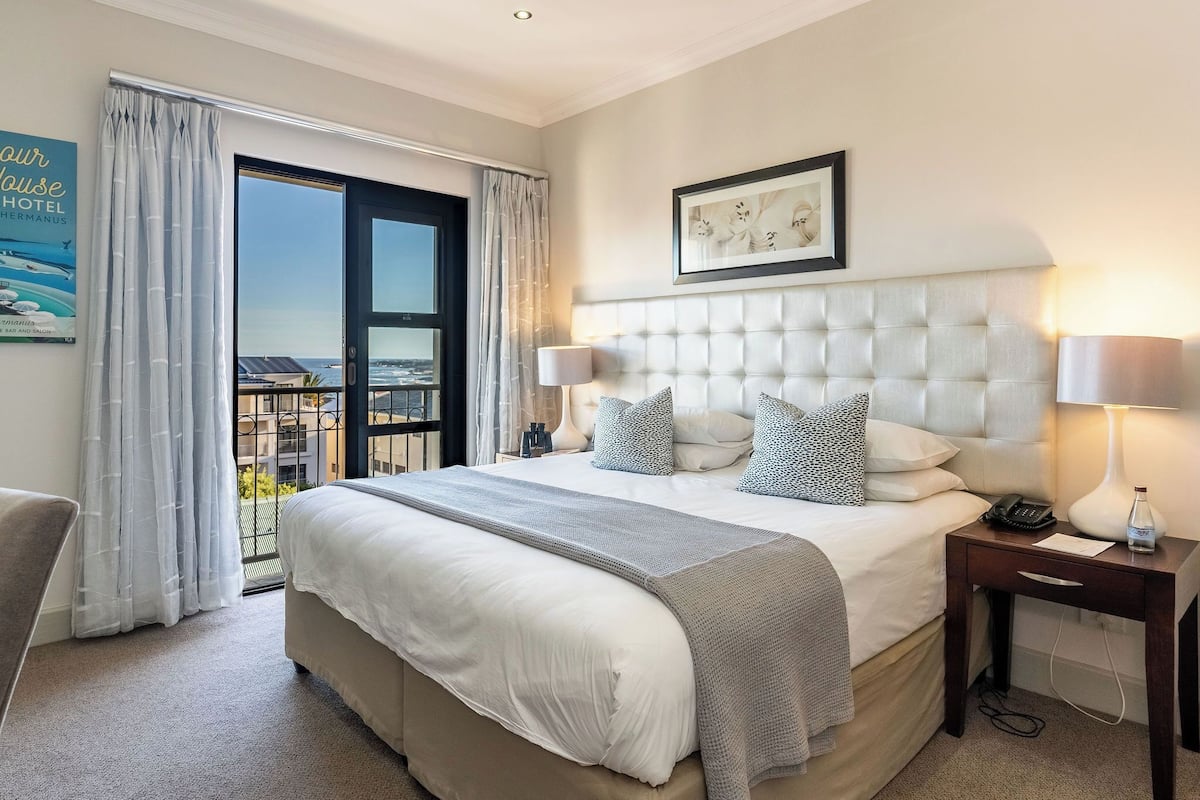 Habitación de hotel con una cama grande, una mesita de noche con lámpara, un cuadro en la pared y vistas a la ciudad desde la ventana.