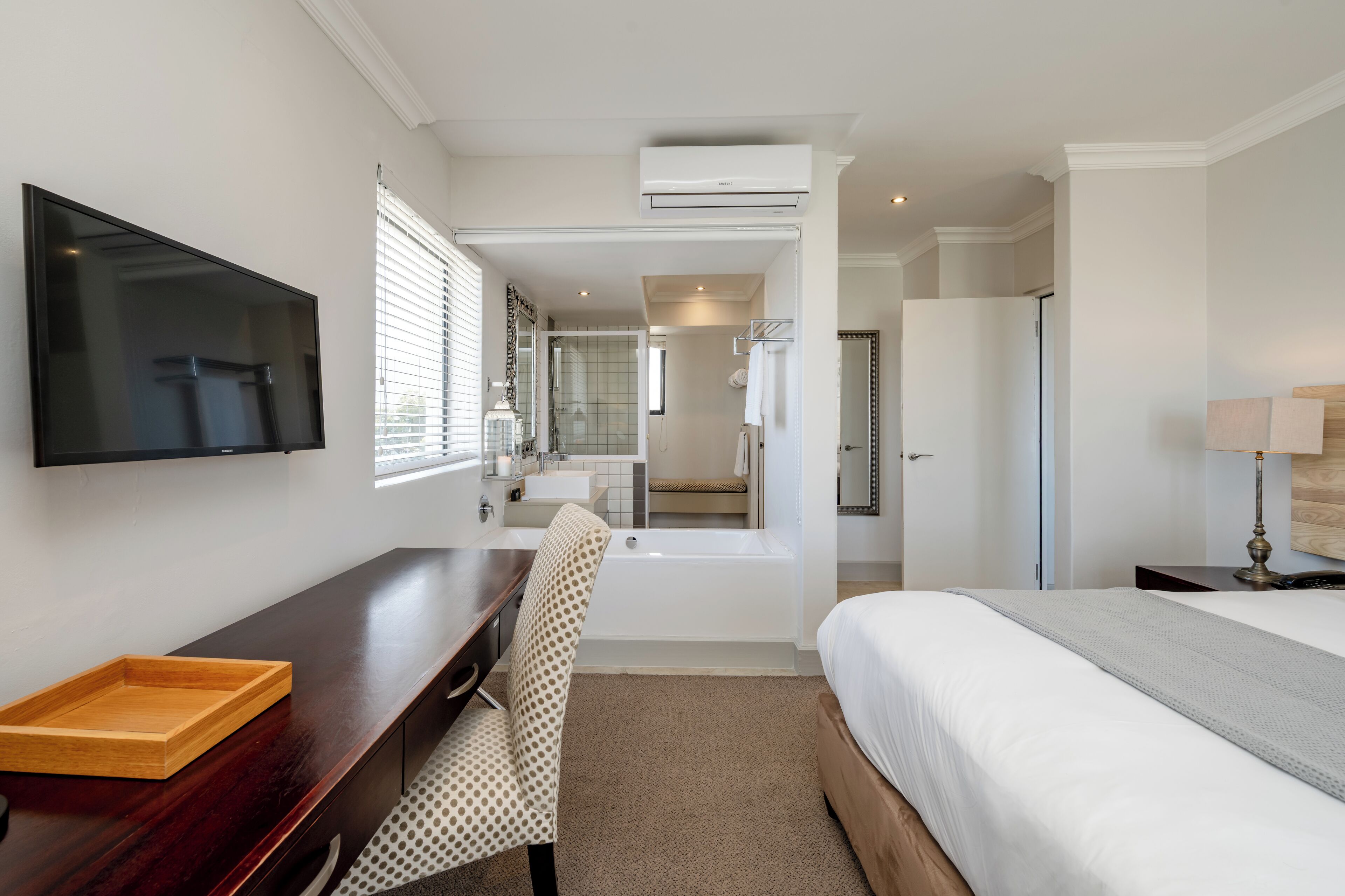 Habitación de hotel con cama, escritorio con bandeja, silla, televisor, bañera y espejo.