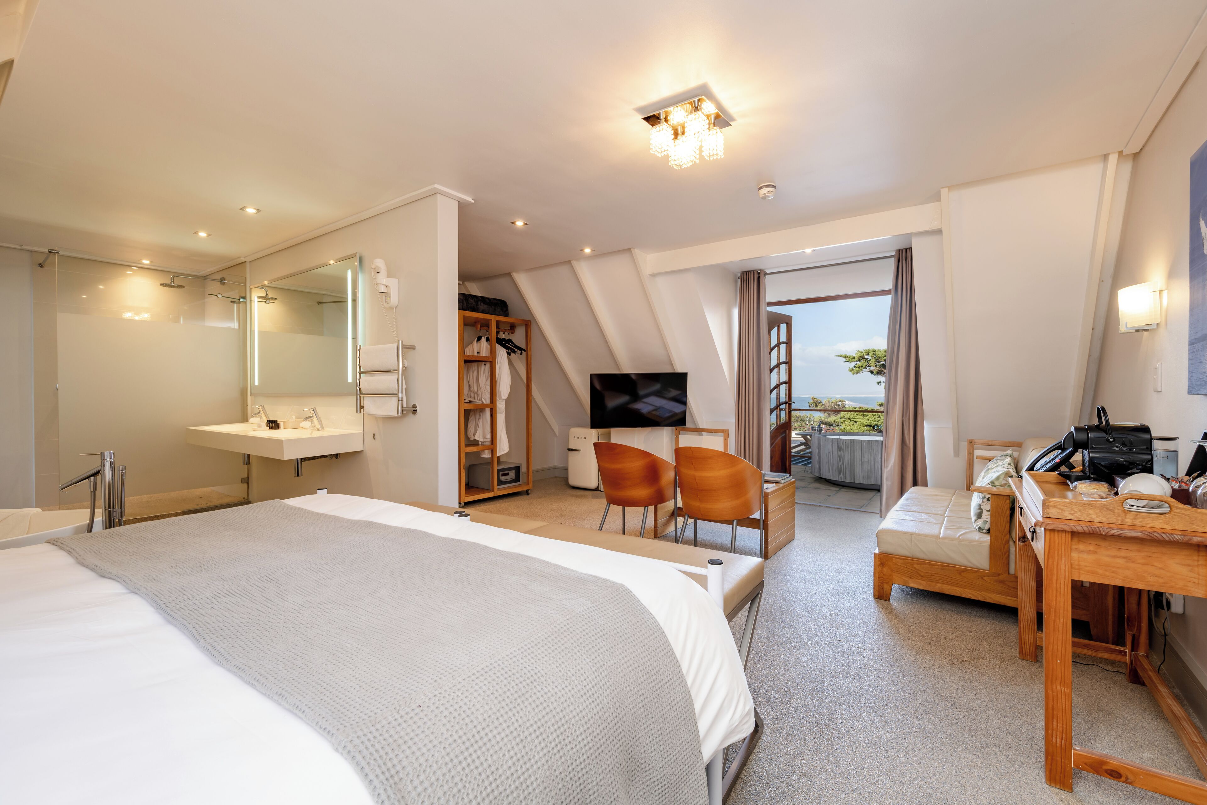 A modern hotel room with a large bed, a desk, a chair, a TV, a wardrobe, and a balcony with a view.