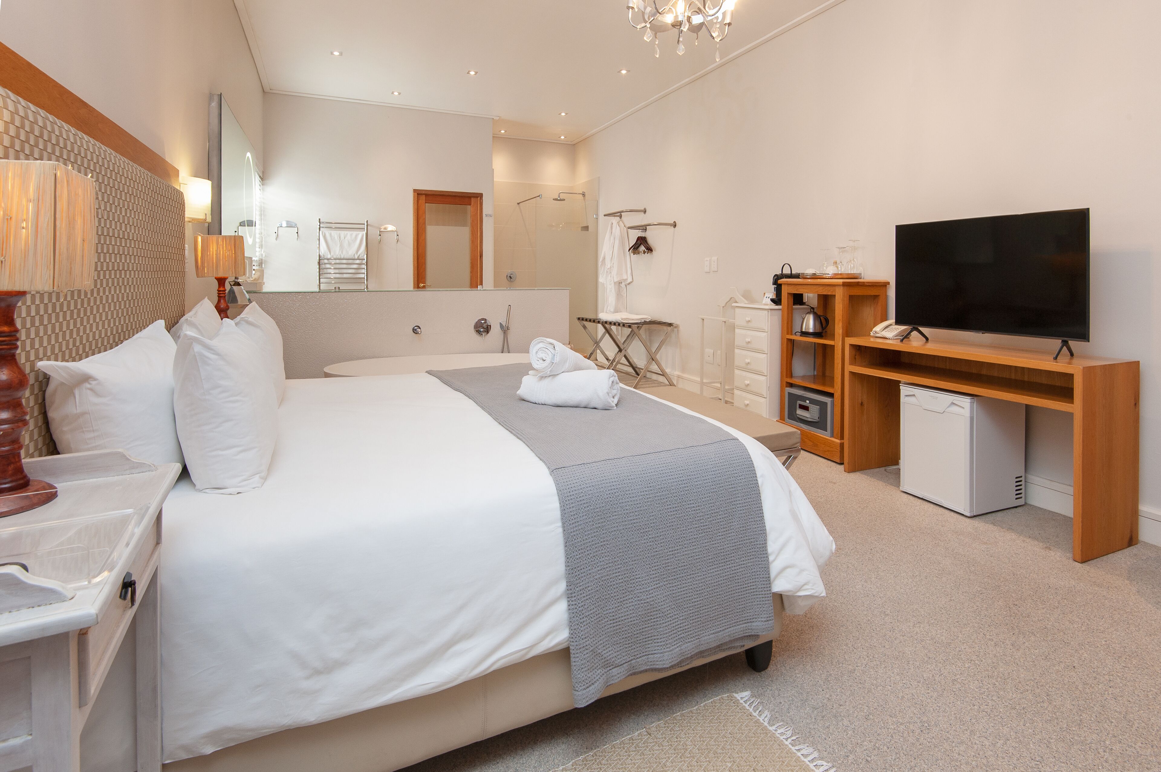 A modern hotel room with a large bed, a flat-screen TV, a desk with a lamp, a mini-fridge, and a shower area.
