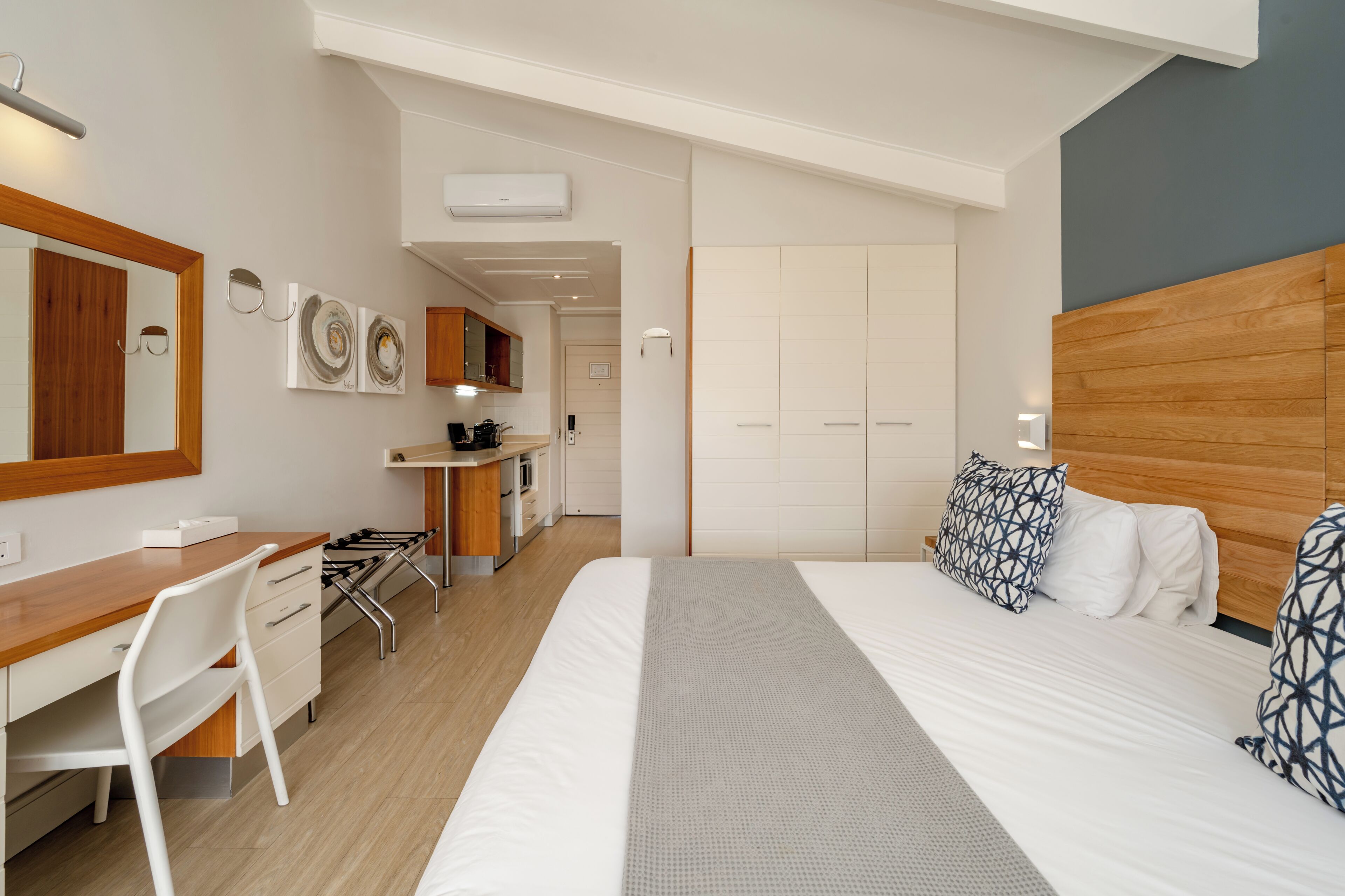 A modern hotel room with a wooden headboard, a desk with a chair, a bed with patterned pillows, and a kitchenette in the background.