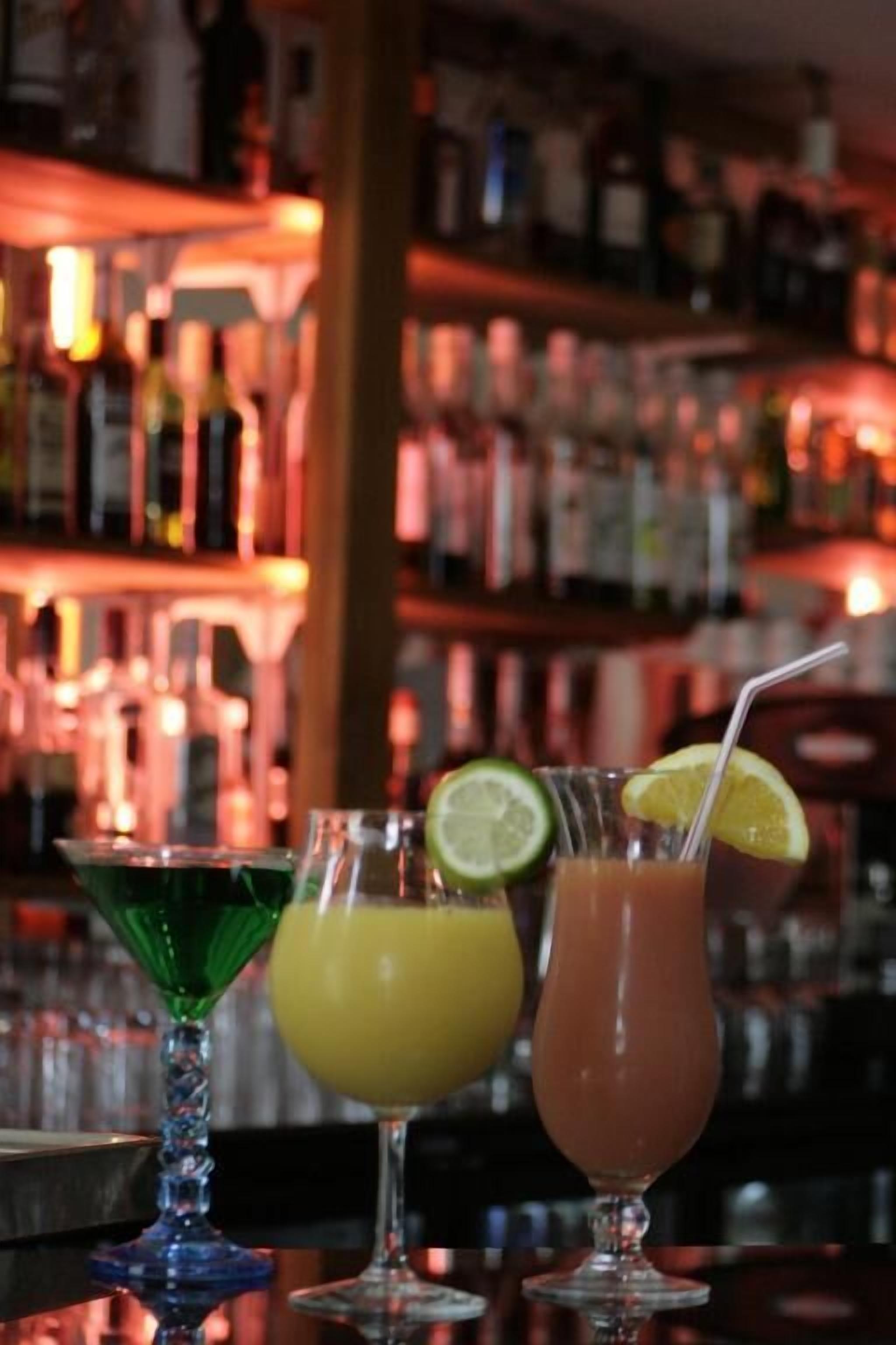 Three cocktails on a bar counter with a blurred background of shelves stocked with bottles.