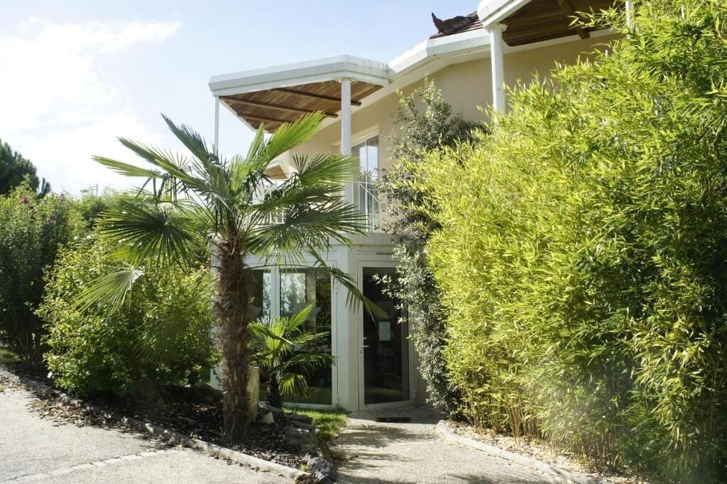 Ein Haus mit überdachter Veranda, Glastür und üppiger, umgebender Bepflanzung.