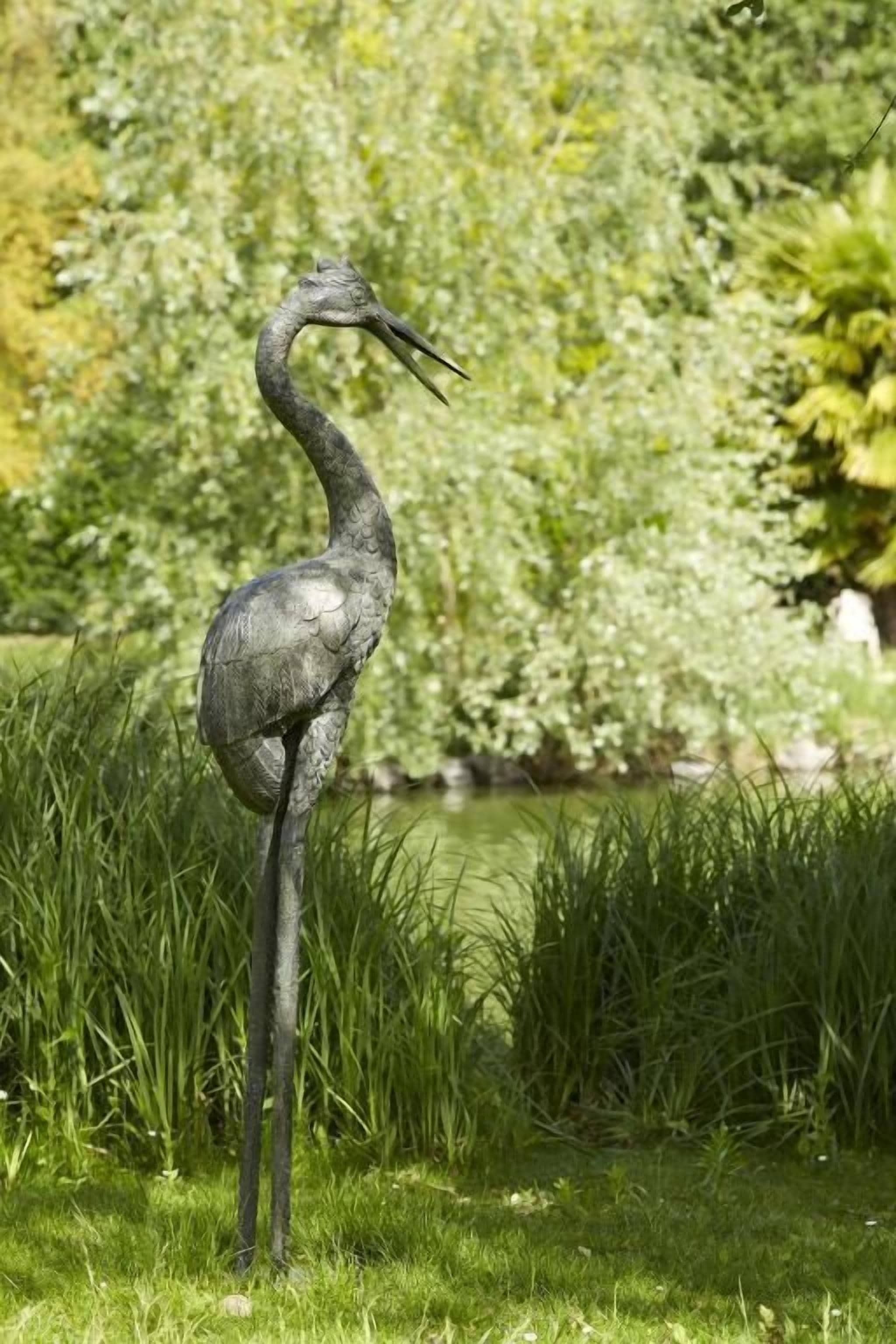 A heron standing in a grassy area with a pond and trees in the background.