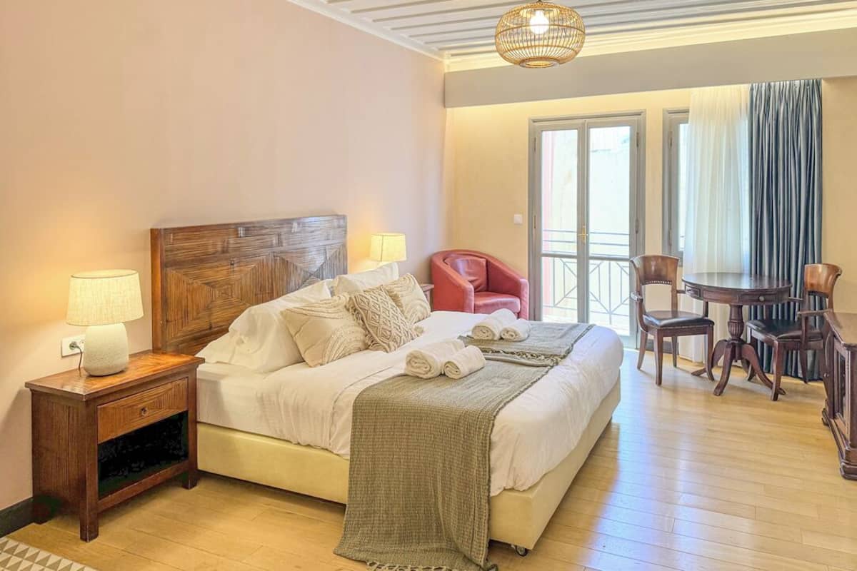 A hotel room with a bed, bedside table, lamp, and a dining area with chairs and a table.