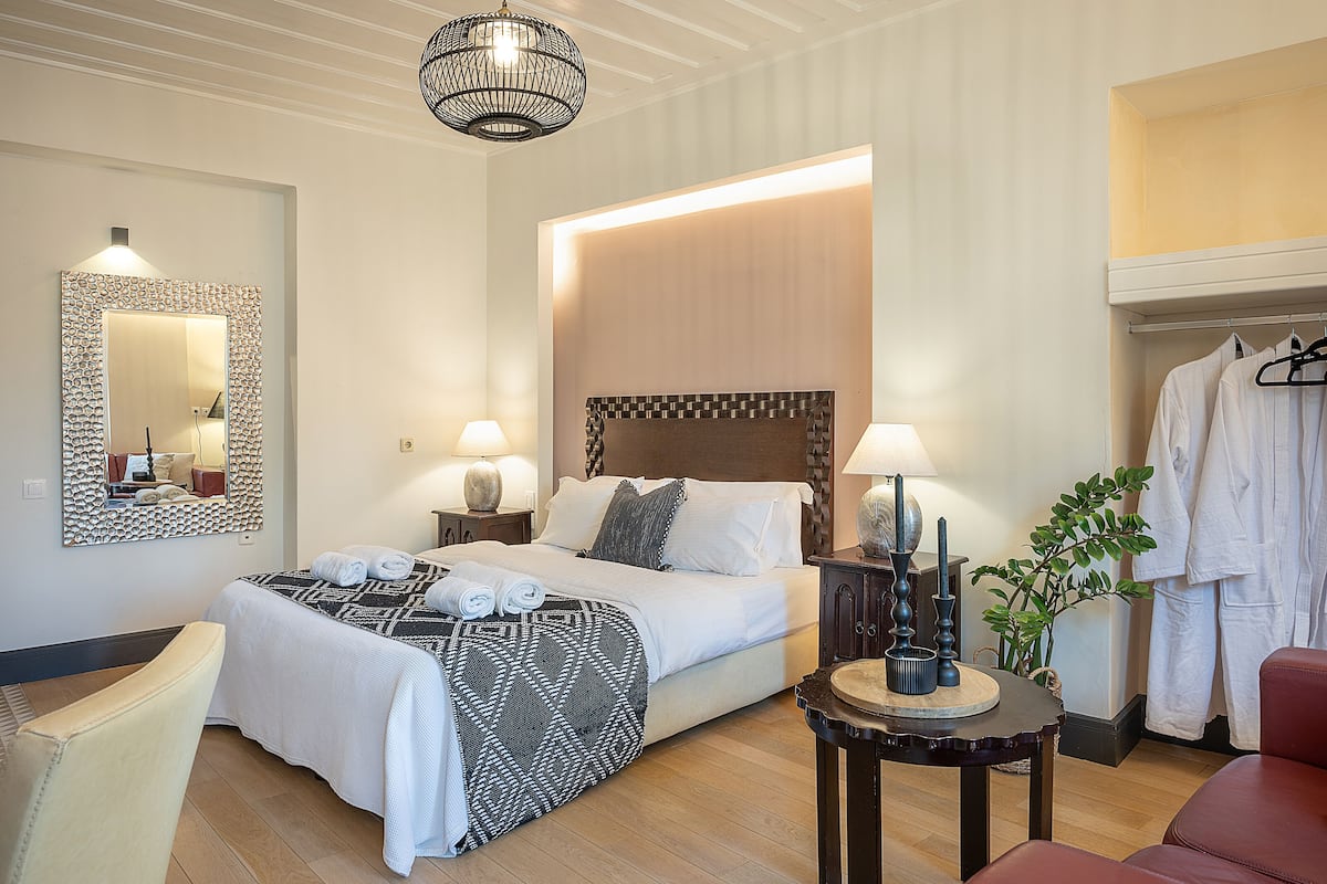 A hotel room with a neatly made bed, two bedside lamps, a round table with a vase, a mirror with a decorative frame, and a coat rack with a robe.