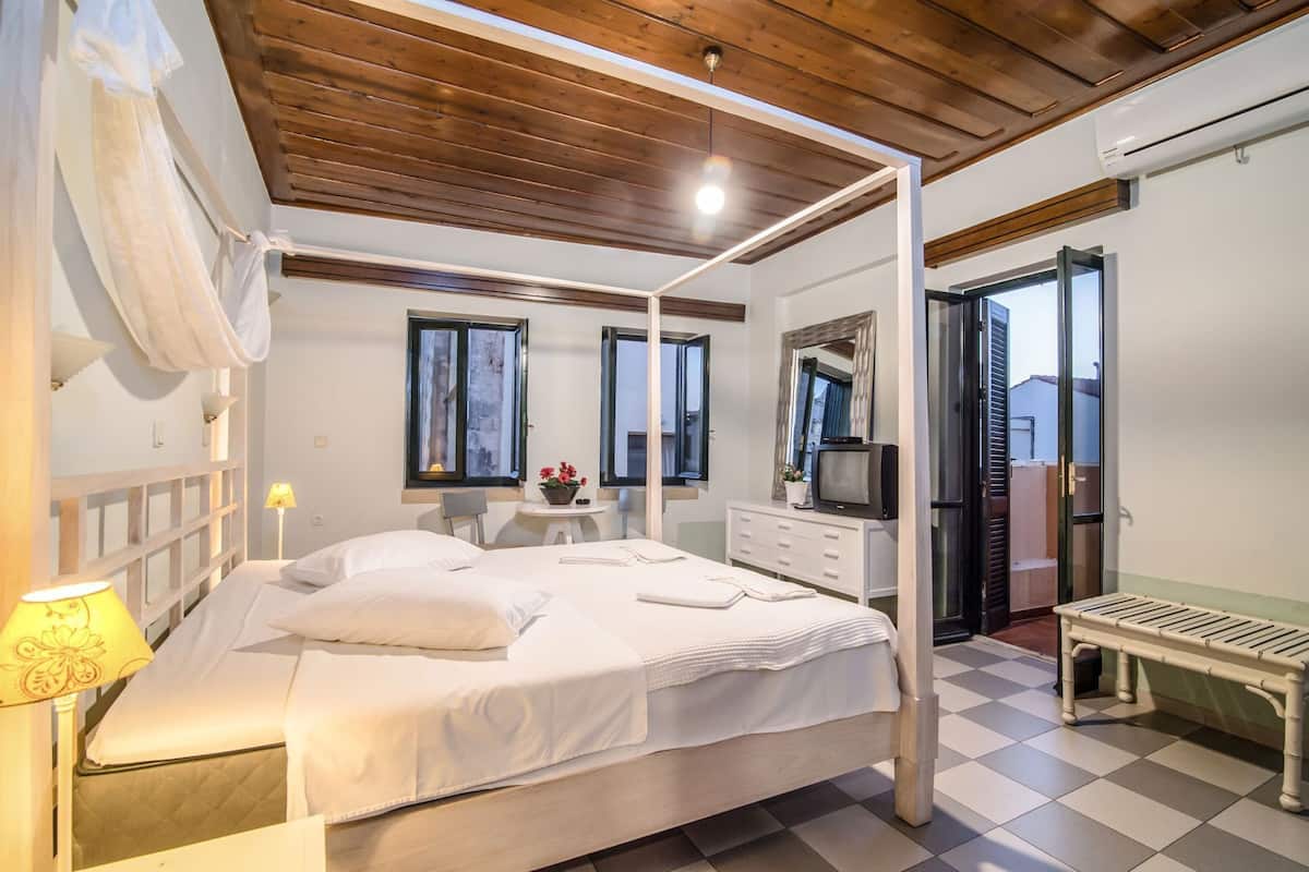A bedroom with a four-poster bed, a bedside table with a lamp, a dresser, a television, and a window with a view.