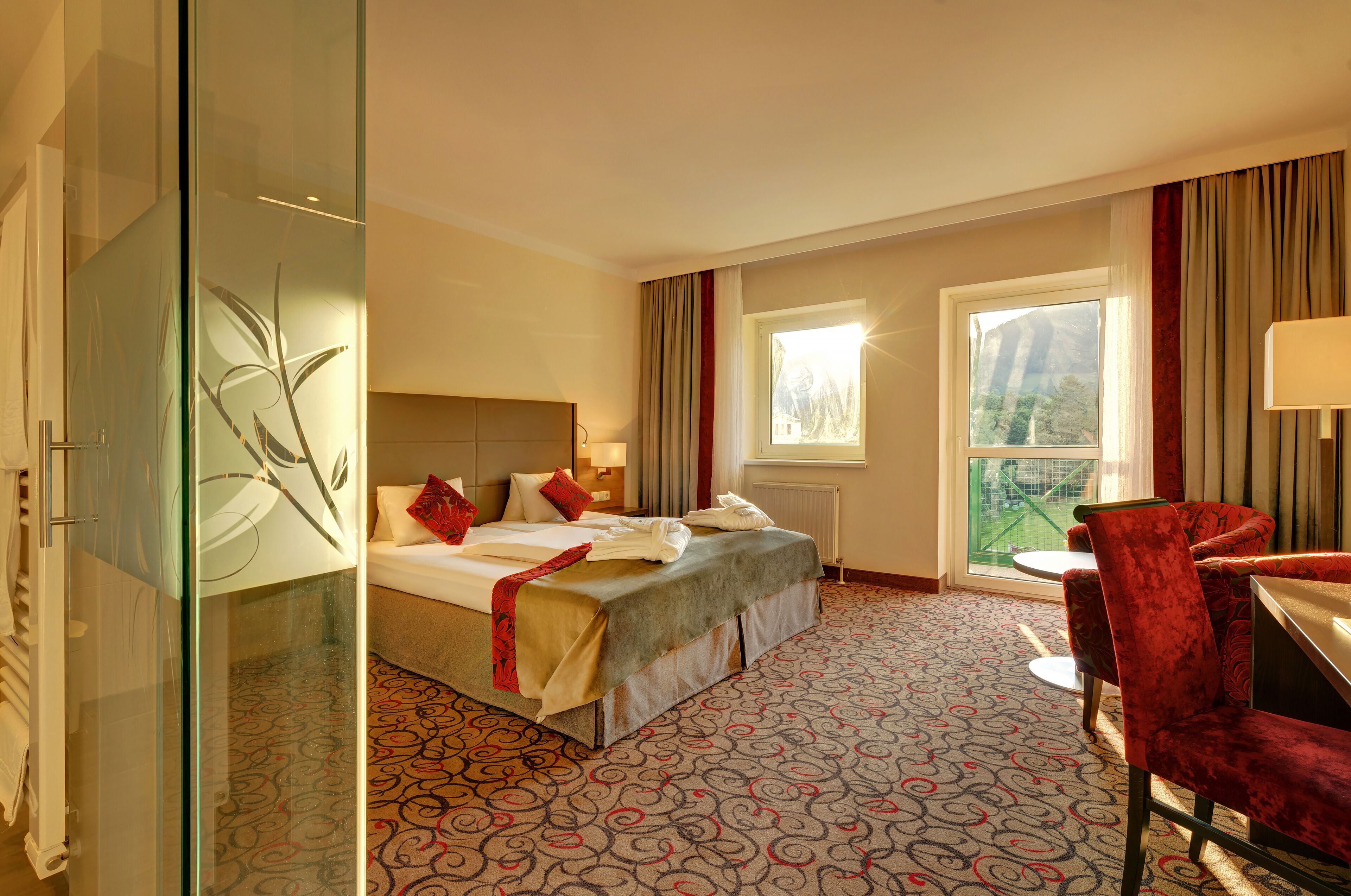 A hotel room with a bed, a desk, a chair, a window, and a glass partition.
