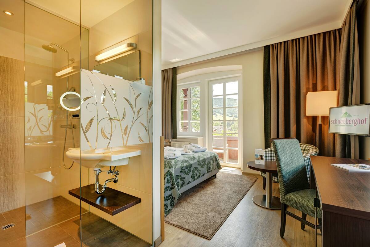 Habitación de hotel con cabina de ducha de vidrio, lavamanos, cama con edredón floral verde, mesa de comedor con una silla y vistas al exterior a través de una ventana.